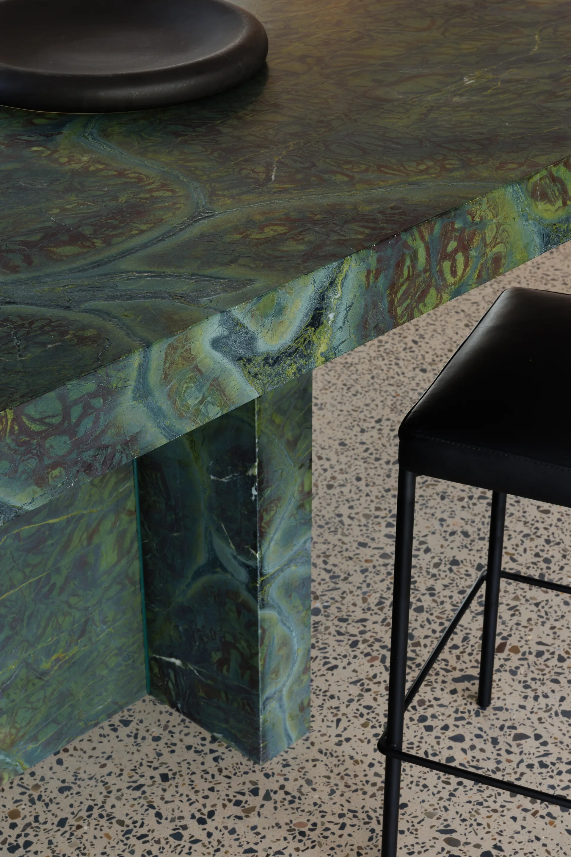 This close-up features a thick, green marble table with intricate swirls of emerald and brown. A black stool with a leather seat stands on the terrazzo floor, which is speckled with small grey and tan stones.
