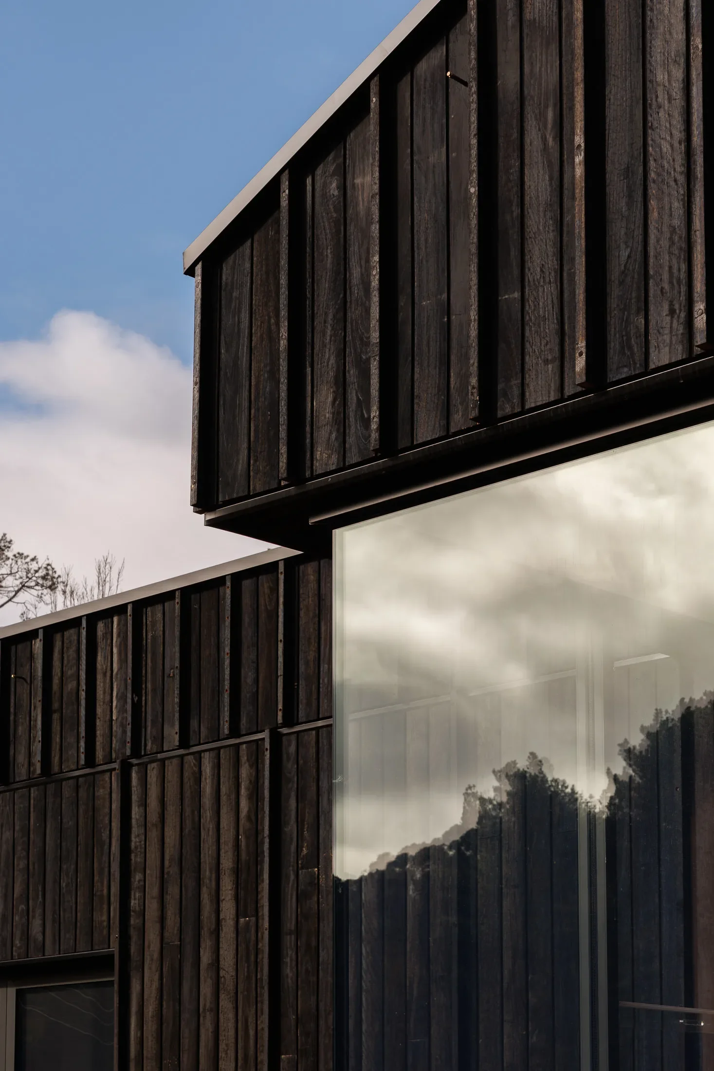 This close-up features the exterior of a modern black building with vertical timber cladding and a clean roofline. A large, floor-to-ceiling glass window reflects the surrounding trees and a cloudy sky, creating a sharp contrast against the dark, textured wood.