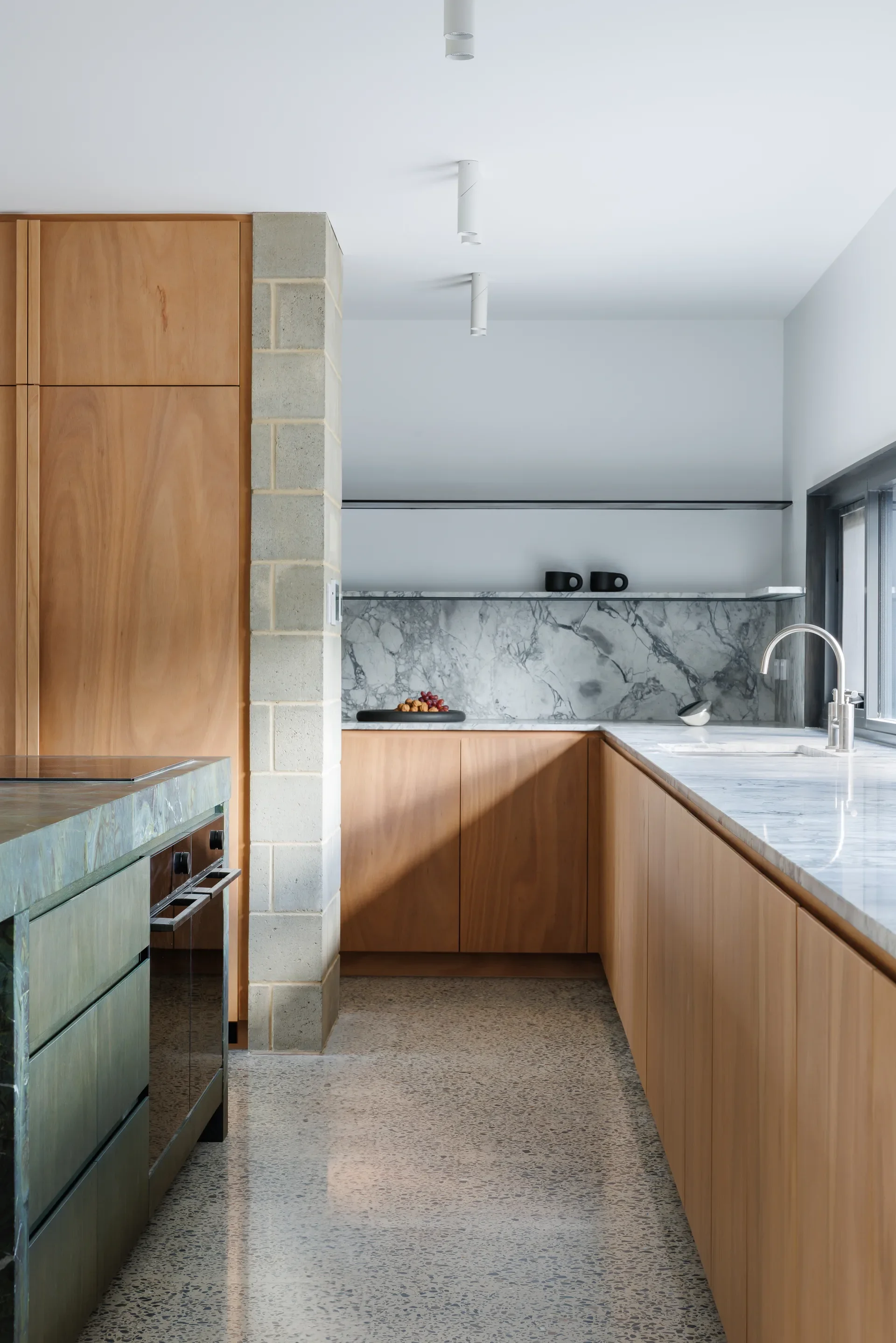 This modern minimalist kitchen features light wood cabinetry, a marble backsplash with gray veining, and sleek white countertops. A built-in sink sits beneath a large window, while open shelving with black mugs and subtle ceiling spotlights enhance the clean, contemporary design.