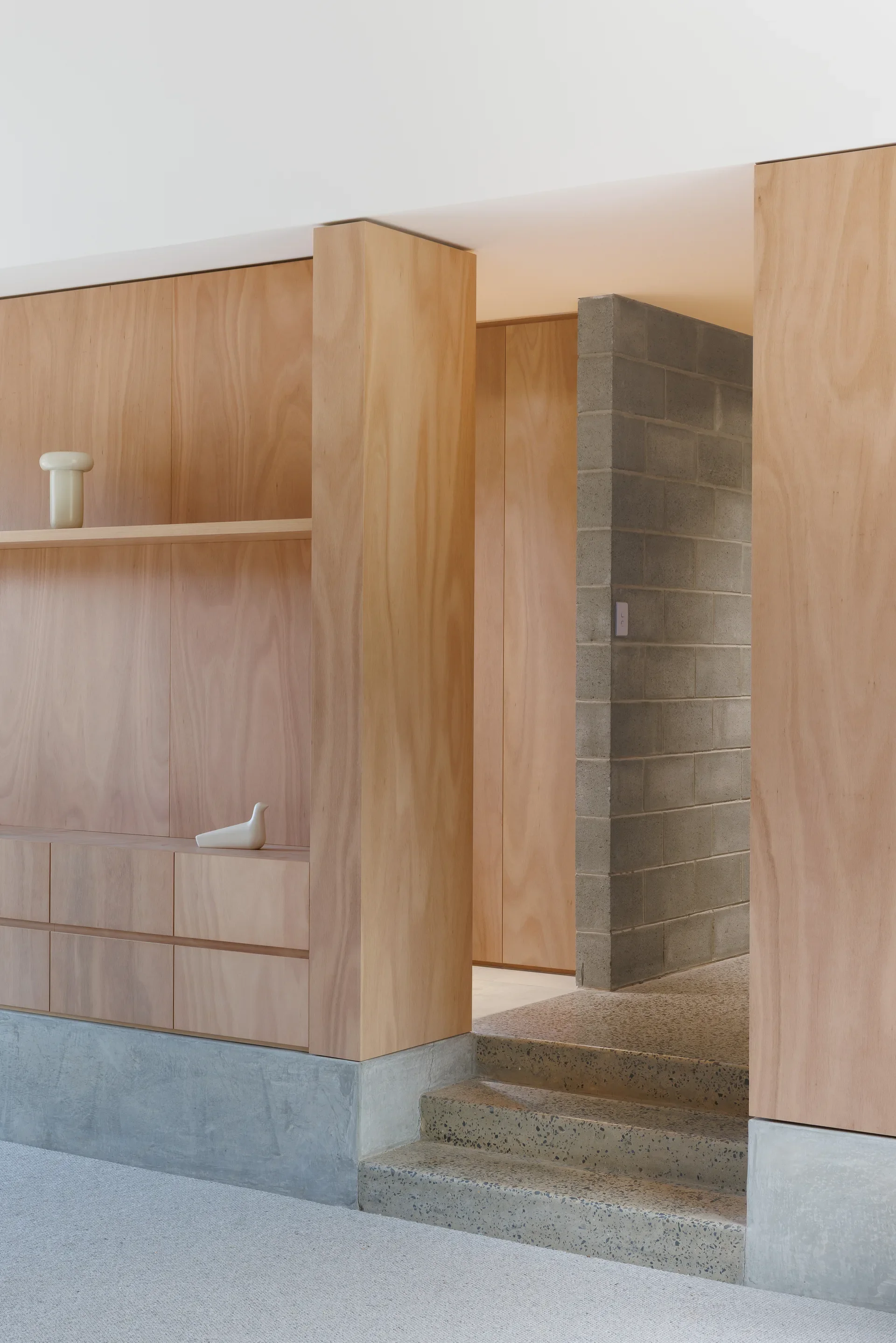 This interior shows light wood built-in cabinetry with open shelving and smooth drawer fronts, accented by a small decorative vase and a sculptural object. A short terrazzo step leads up to a hallway framed by a gray concrete block wall, with polished concrete and terrazzo surfaces adding texture to the warm, natural wood tones.
