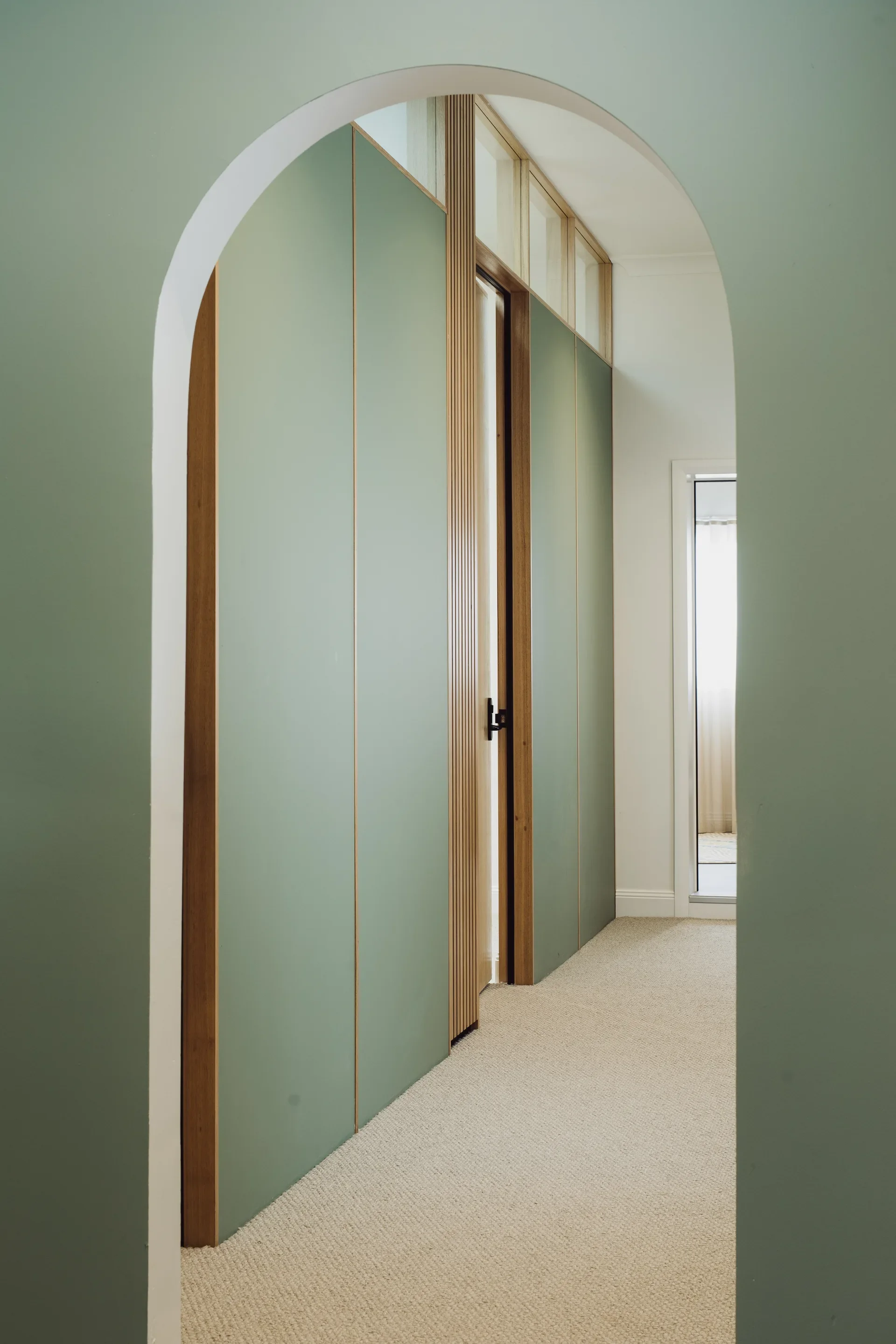 A hallway framed by a sage green arched doorway. The corridor features light green cabinetry with timber trim, a slatted wood accent, and neutral textured carpeting, creating a calm, cohesive aesthetic.