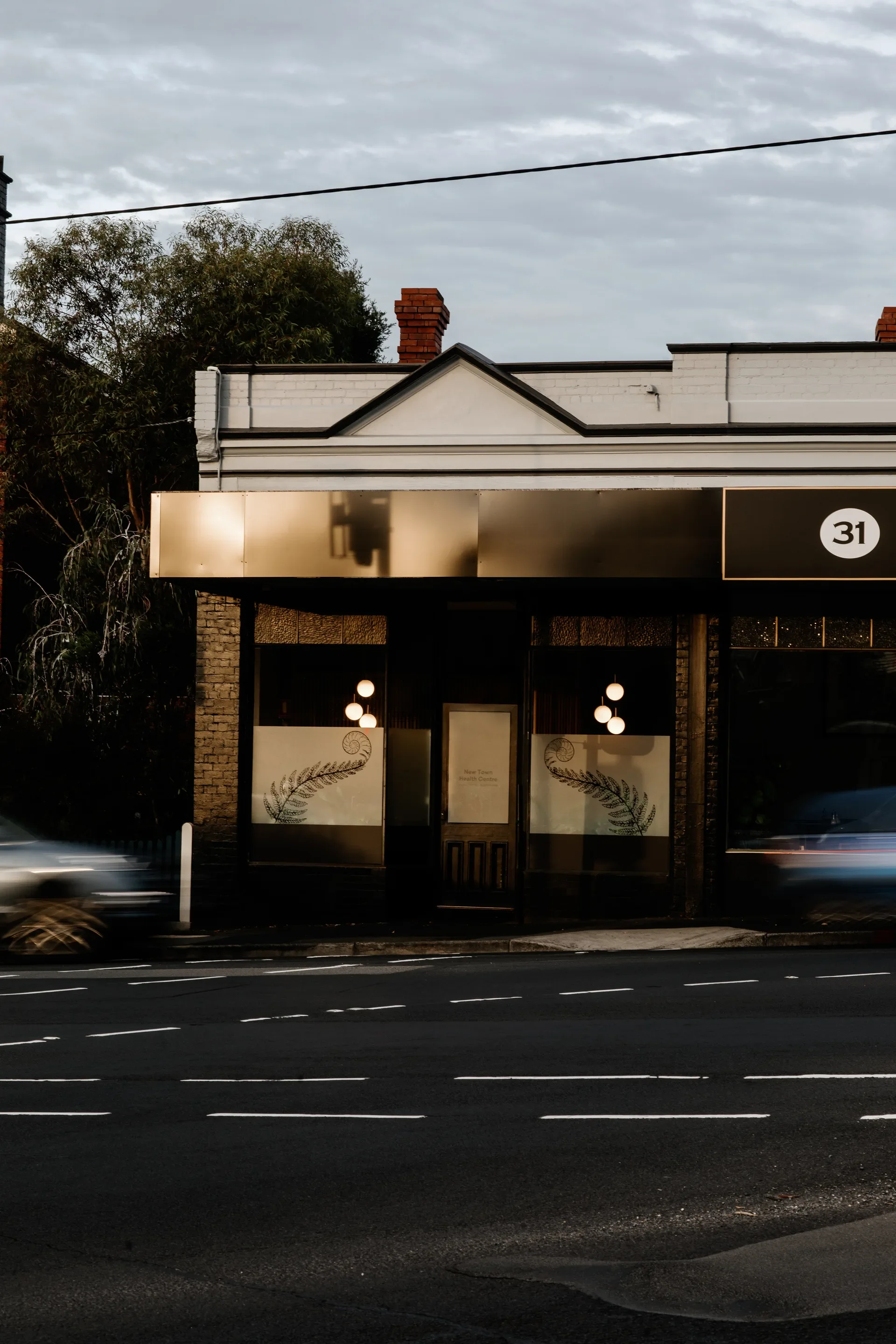 A storefront at dusk with a gold-metallic awning reflecting the dim sky. The windows are decorated with large fern illustrations and illuminated from within by warm globe pendants, while the blurred motion of passing cars creates a moody, urban atmosphere.
