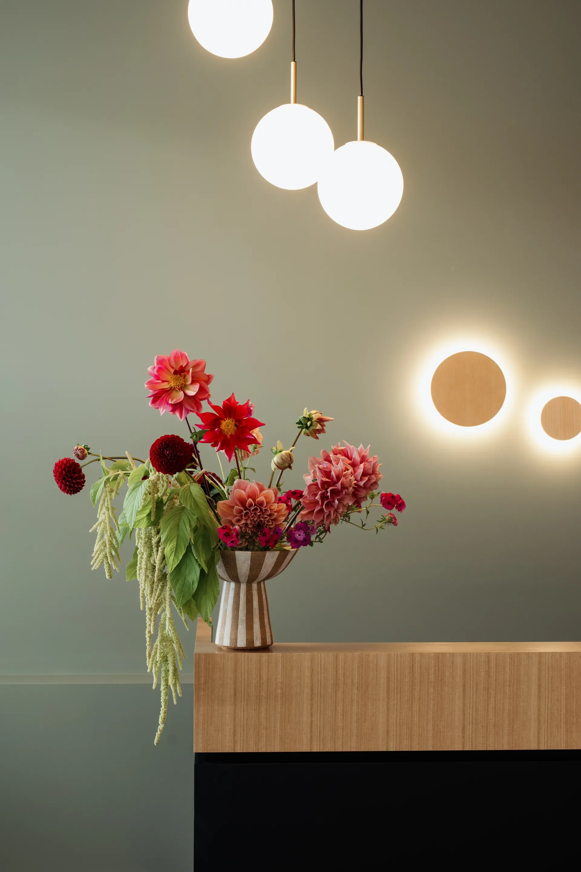 A close-up of a reception desk featuring a vibrant floral arrangement in a striped vase. The scene is set against a sage green wall, illuminated by hanging white globe pendants and two circular, glowing wall sconces.