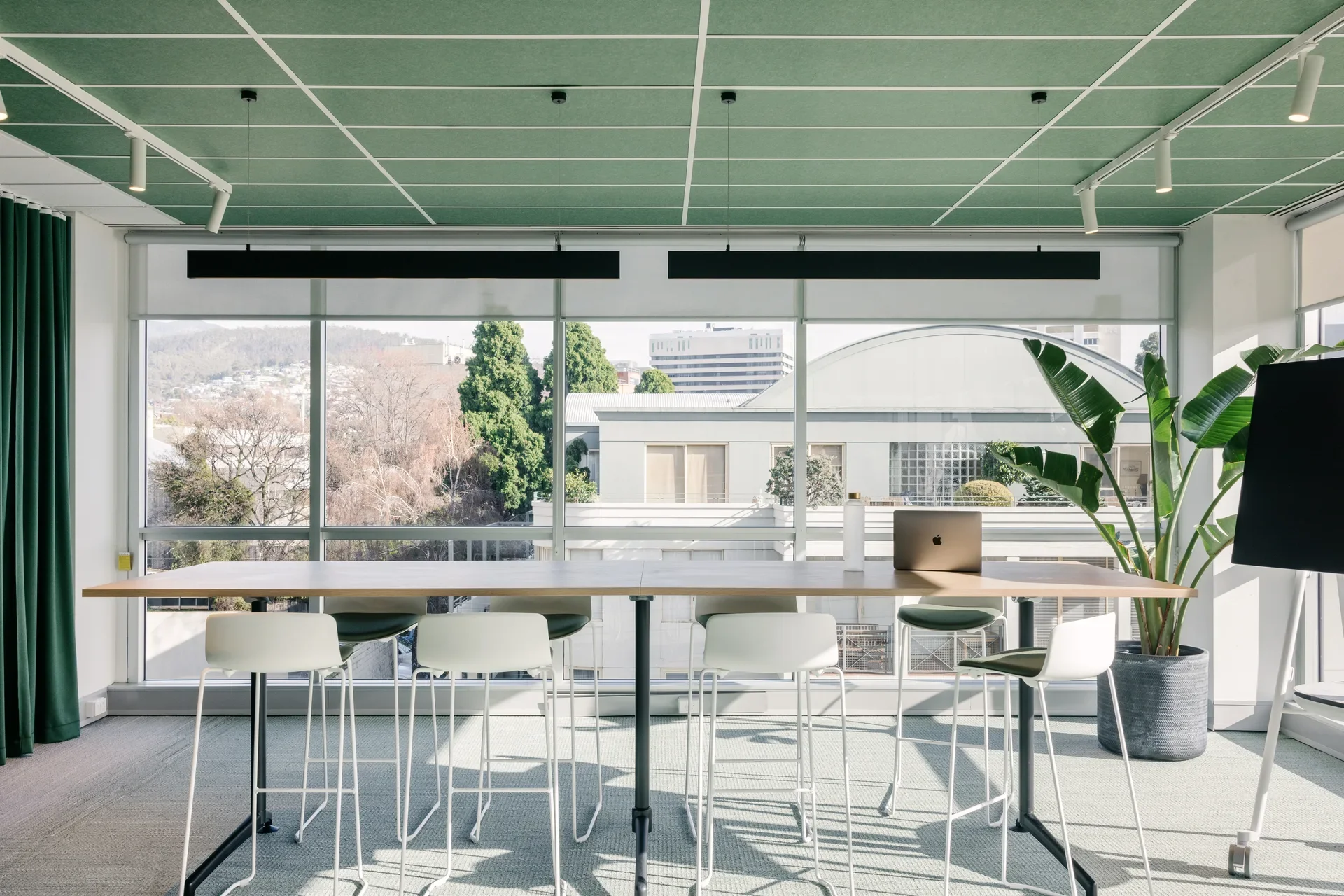 Gemini said This bright workspace features a long wooden table with white barstools and a laptop. Large windows offer a city view, complemented by a green-tiled ceiling and a large potted plant.