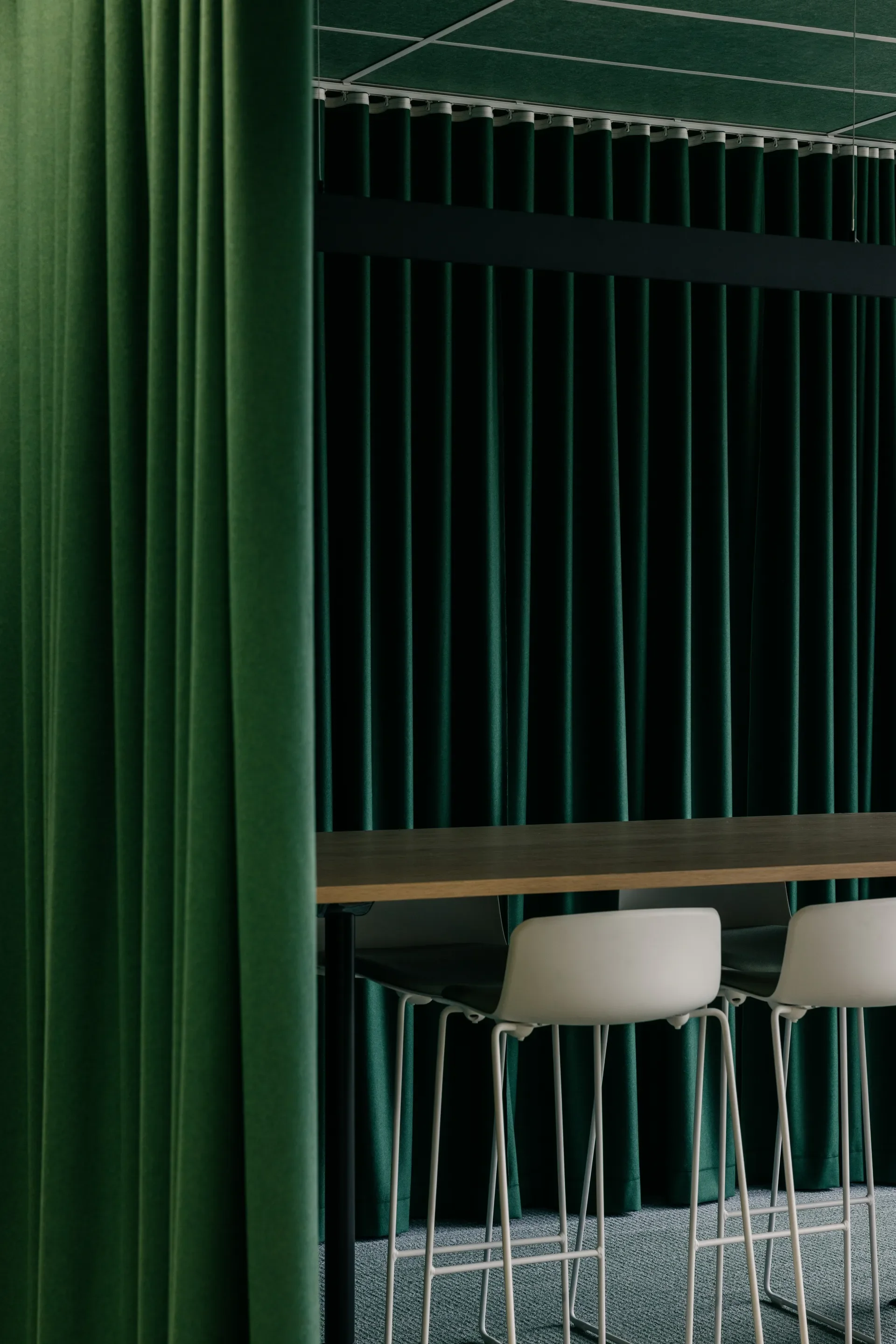 This modern meeting area features white high-top chairs at a wooden table, set against a backdrop of floor-to-ceiling green curtains. The space is finished with a matching green-tiled ceiling and dark track lighting.