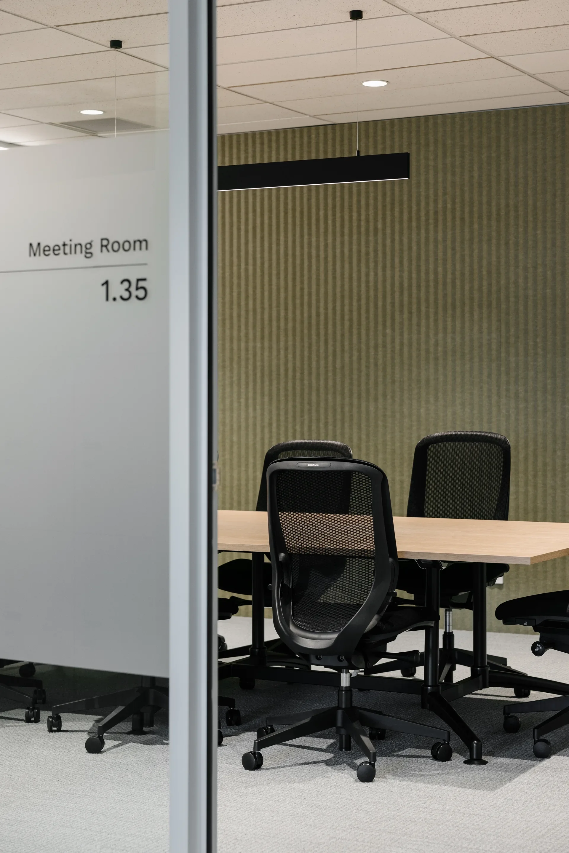 A wooden table with black rolling mesh chairs are set in a meeting room. The space features a textured green wall and a black pendant light.