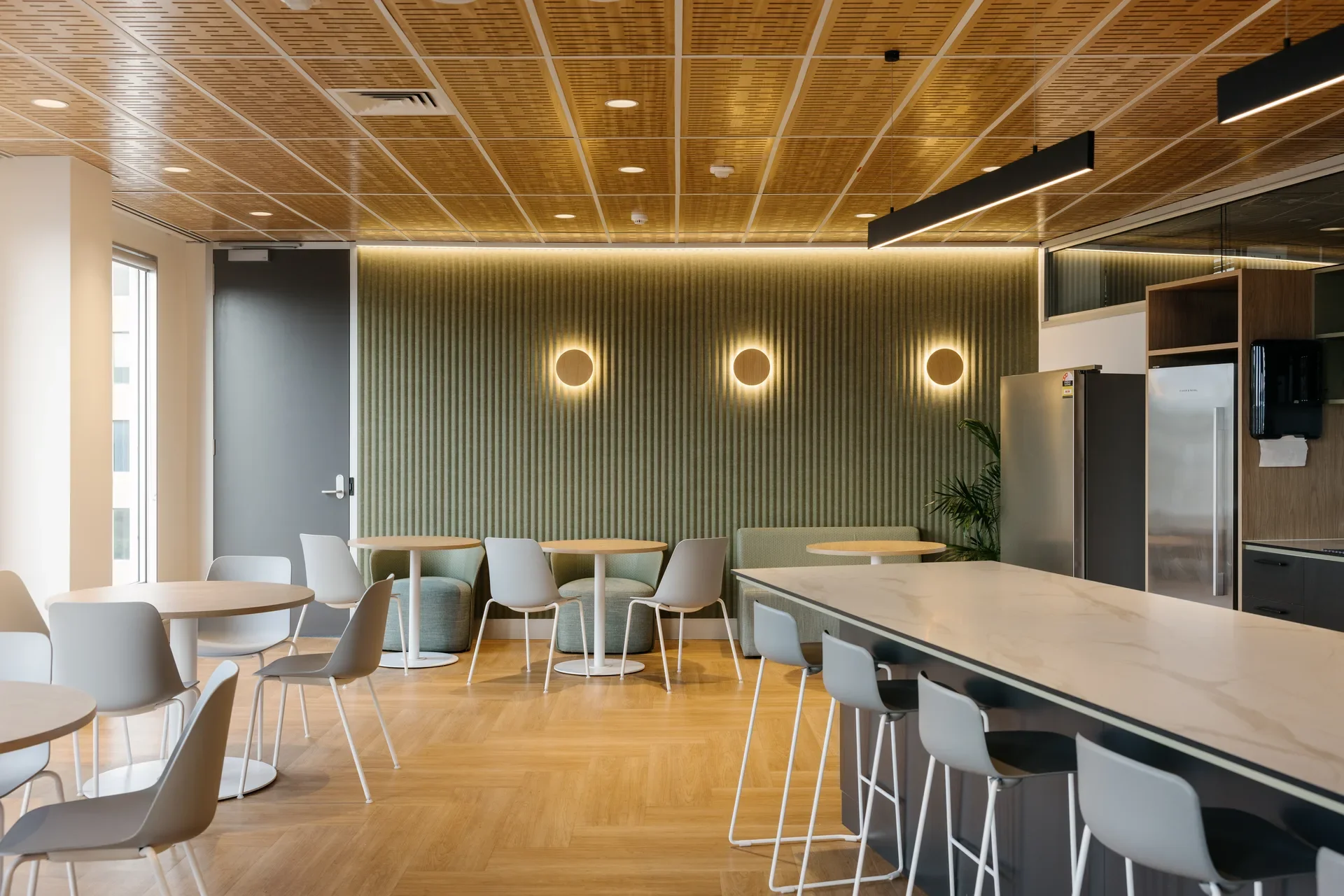 This staff kitchen features vertical-slat wood walls accented by three circular wall sconces and a warm LED light strip. The space is furnished with white and grey chairs at small round tables, beneath a perforated wood-panelled ceiling.