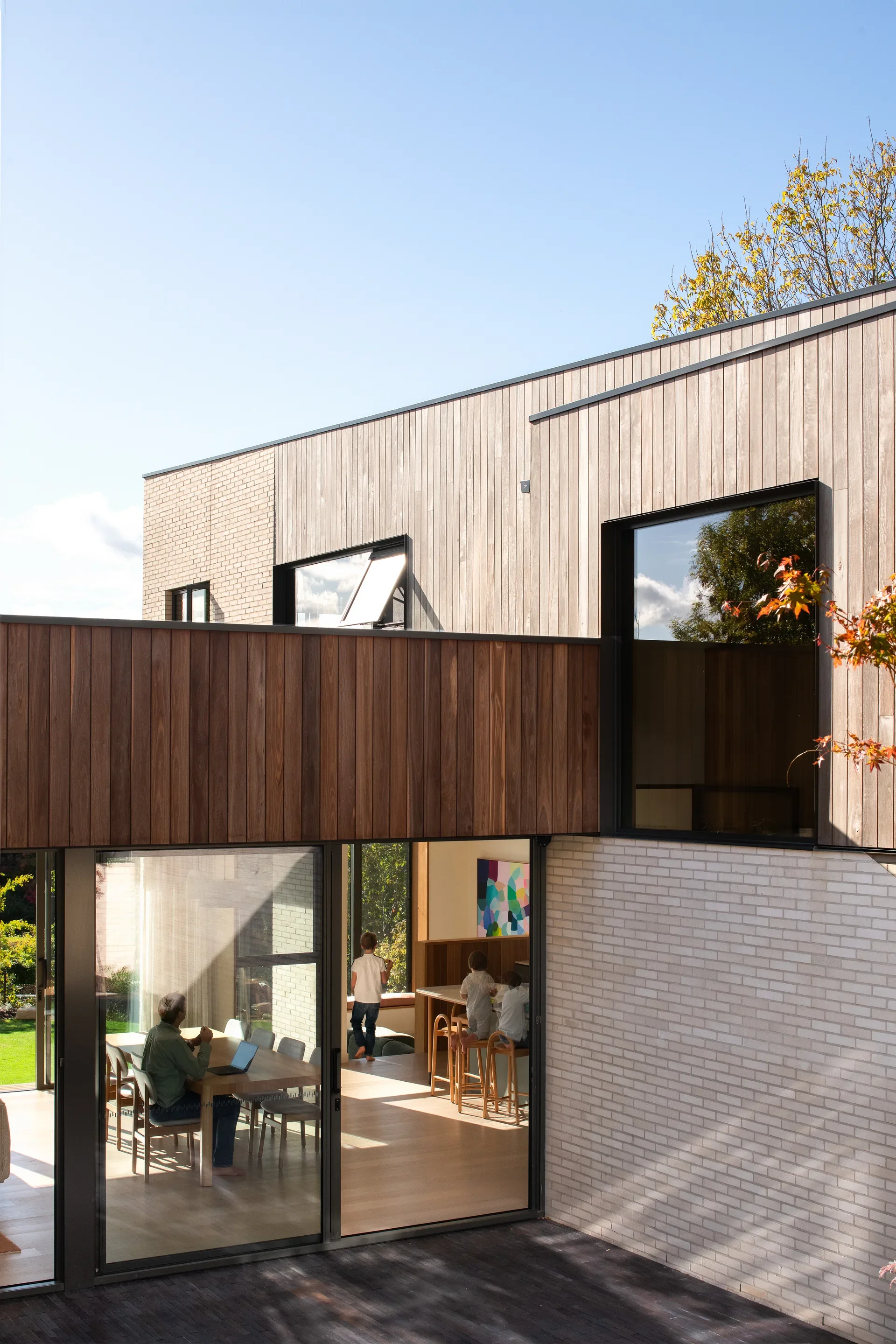 This exterior view shows a two-story home featuring a combination of vertical wood cladding and white brick. Large glass doors reveal a well-lit dining and kitchen area where a person works at a table and children sit at an island.