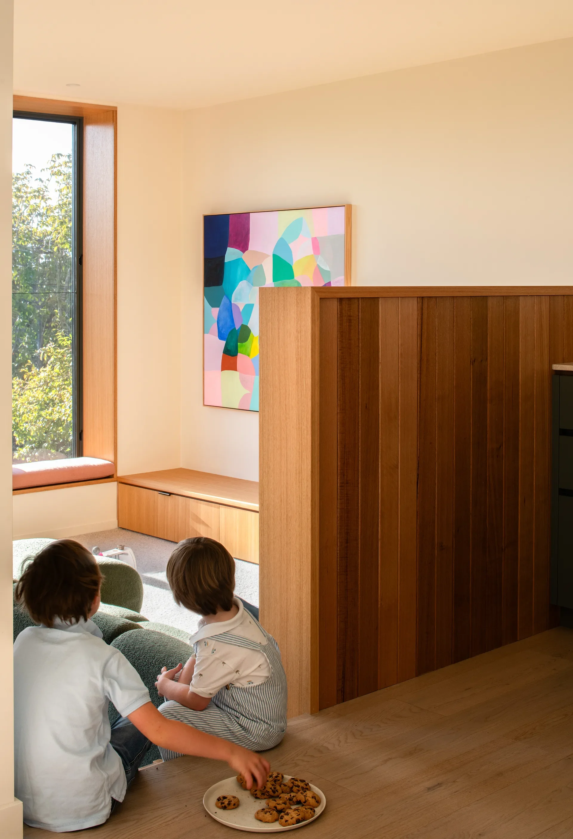This sunlit interior features two children sitting on a carpeted floor next to a plate of cookies and a wooden partition. A vibrant, abstract painting hangs on the white wall near a window seat that overlooks lush green trees.