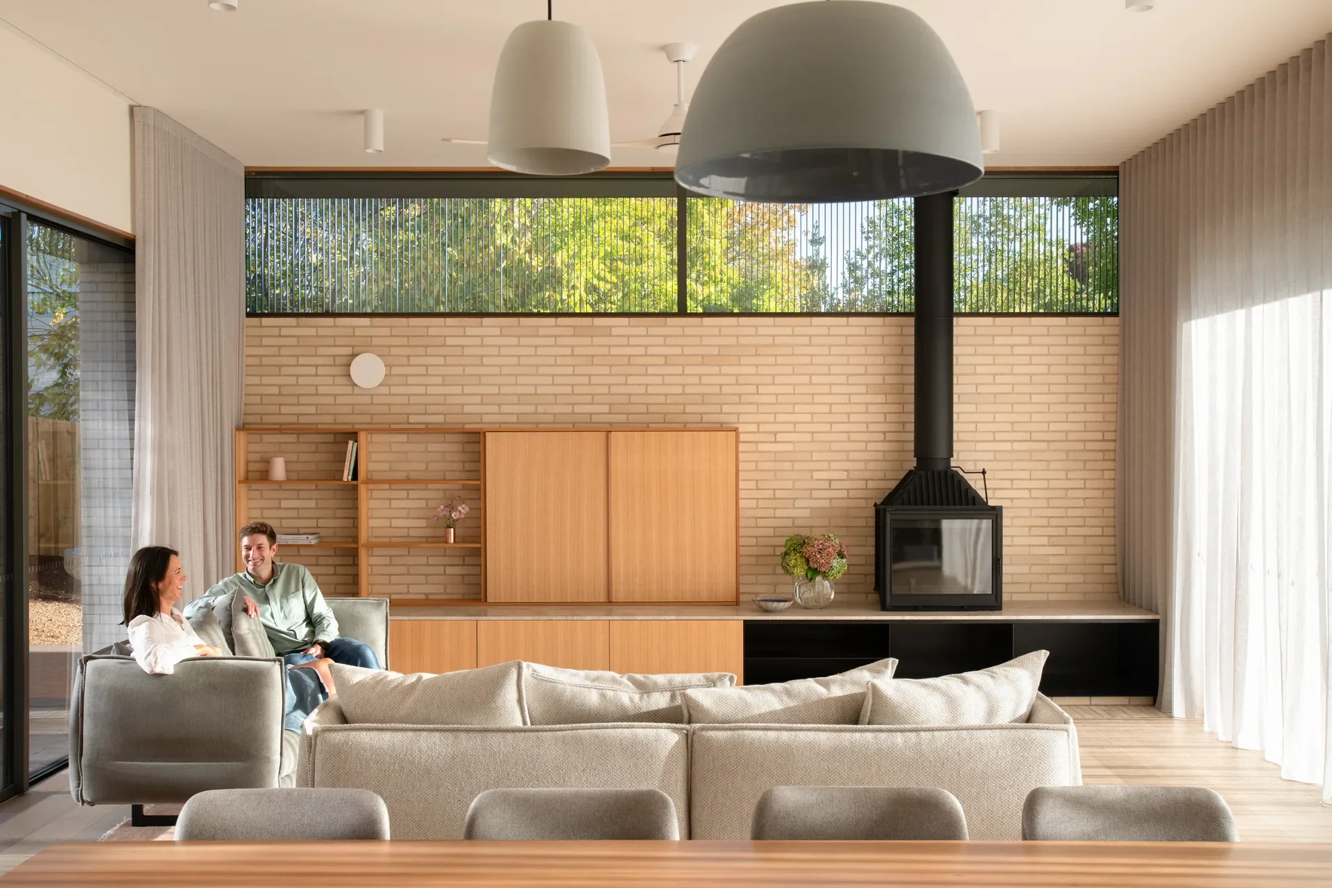 The living room features a black fire place set against a light brick wall and a long wooden storage unit. A couple relaxes on a grey sofa, while large dome-shaped pendant lights hang from the ceiling in front of a wide window with sheer curtains.