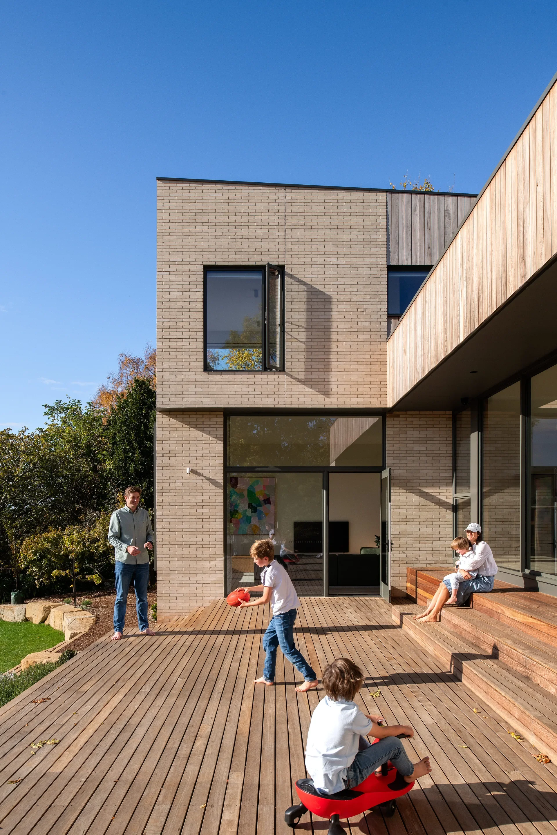 This exterior view shows the two-story home with a mix of light brick and vertical timber cladding. A family is enjoying the spacious wooden deck, where children play with a ball and a ride-on toy under a clear blue sky.