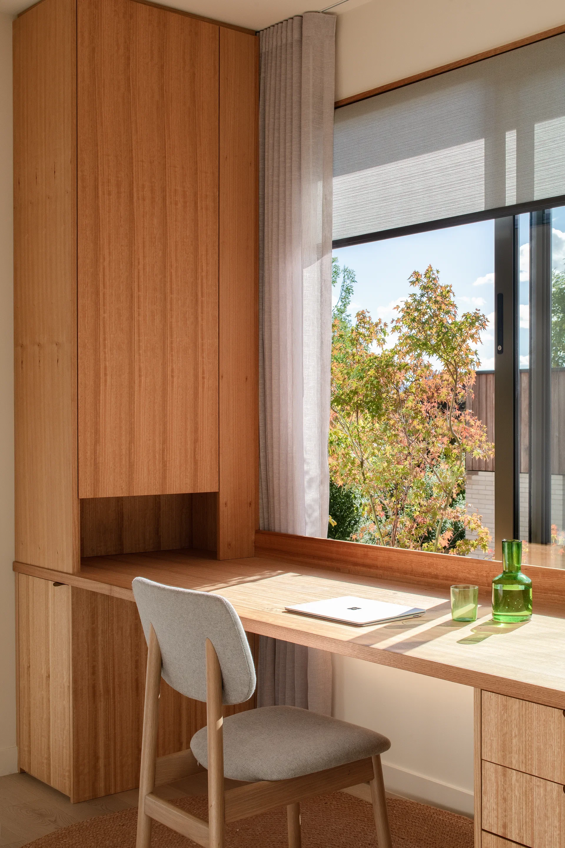 This sunlit workspace features a built-in wooden desk and a tall storage cabinet placed next to a large window. A grey upholstered chair sits at the desk, which overlooks a view of autumnal trees and a neighbouring building under a clear sky.