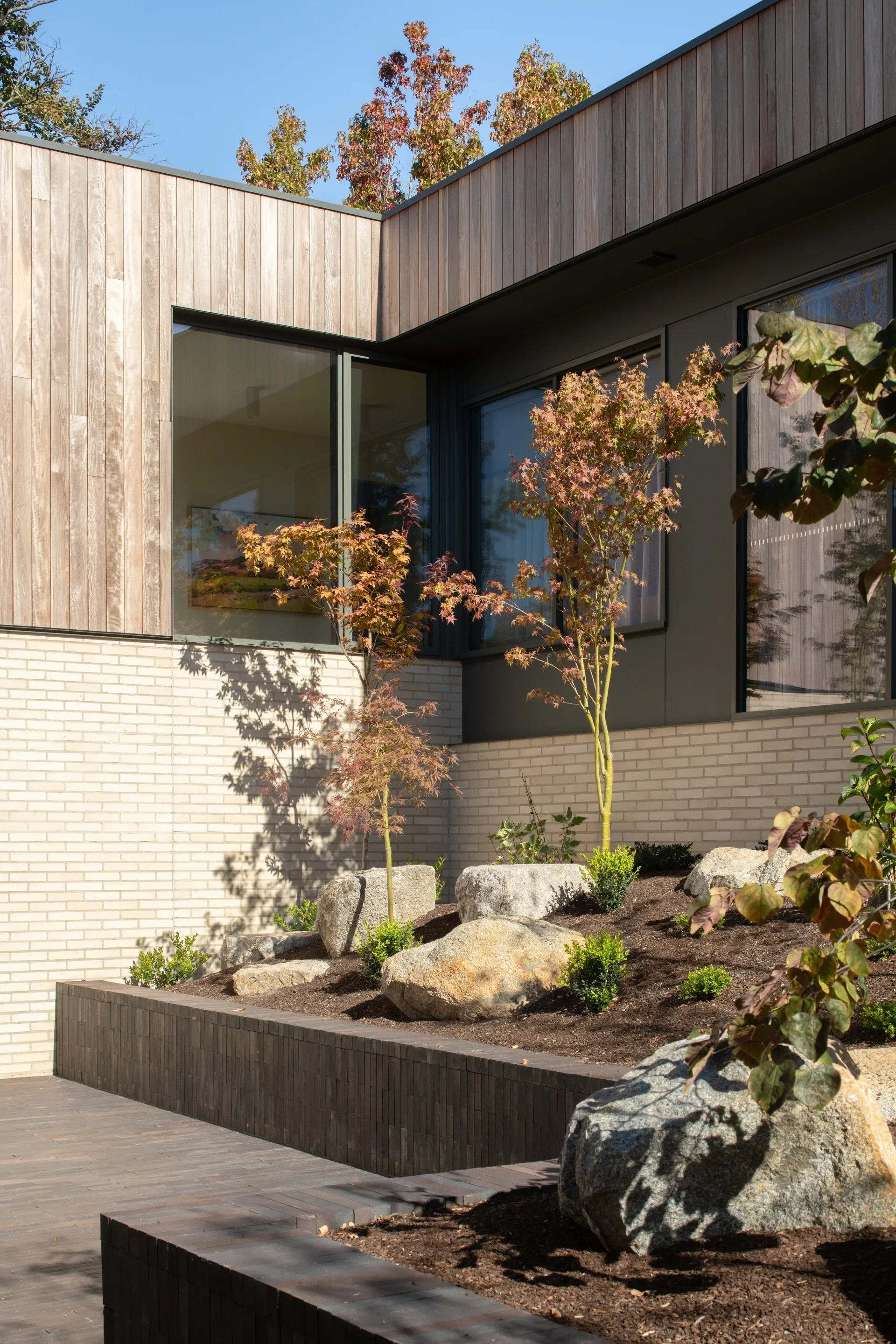 This outdoor courtyard features a sloped garden bed with boulders and young trees, situated between a white brick wall and a dark wood-panelled upper story. A dark-toned retaining wall separates the garden from a paved patio area under a clear sky.