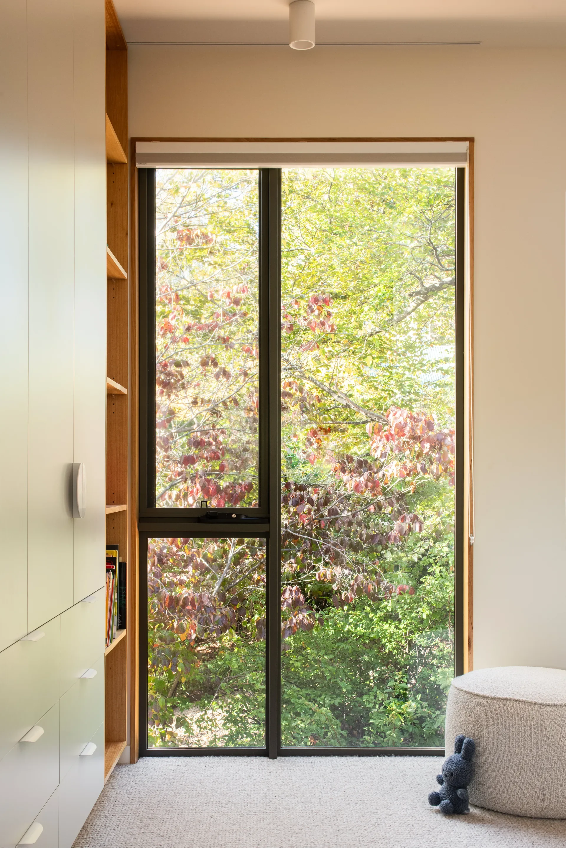 This interior features a tall window framing a lush view of green and reddish trees. A white storage unit and a round ottoman sit on the neutral carpet, creating a minimalist, sunlit space.