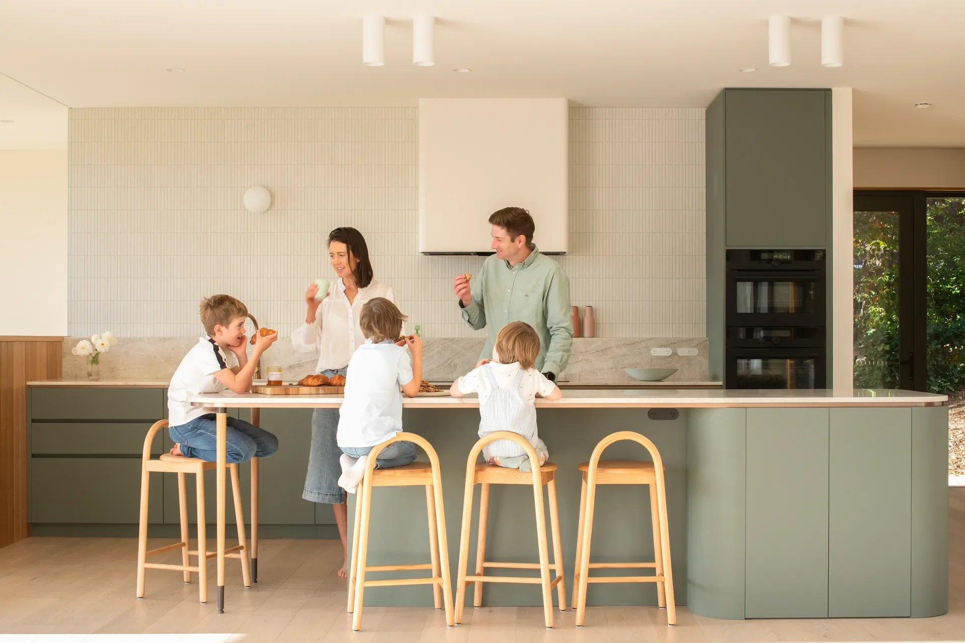 This modern kitchen features a long sage-green island where a family is eating breakfast. The space is finished with light wood floors, a vertical tiled backsplash, and minimalist white cylindrical lights on the ceiling.
