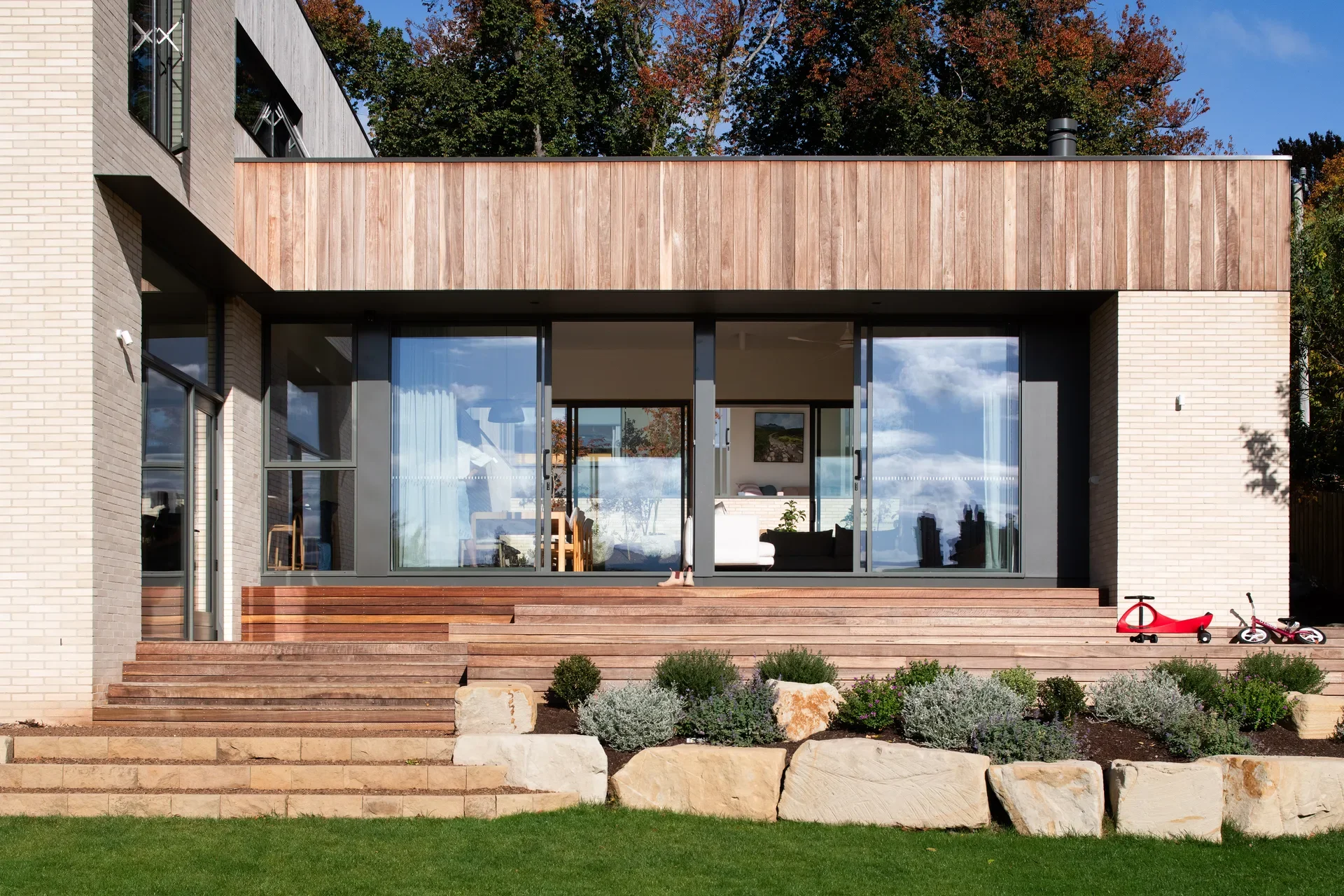 This exterior view shows a two-story home combining light brickwork with vertical timber cladding. Wide wooden steps lead from a grassy lawn, past a stone-bordered garden bed, to a large glass sliding door that reveals the interior living space.