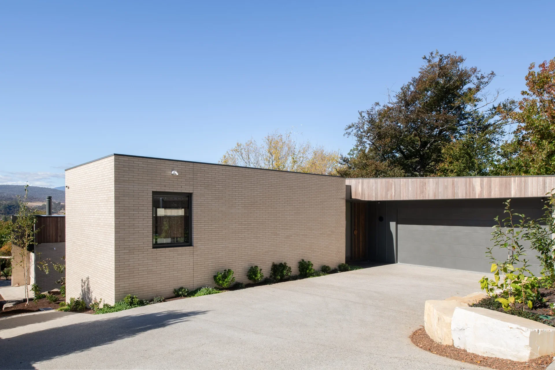 This exterior view features a low-profile home with a light tan brick facade and a flat roofline. A wide concrete driveway leads to a dark grey double garage, while minimalist landscaping with small green shrubs and large white stones borders the property.