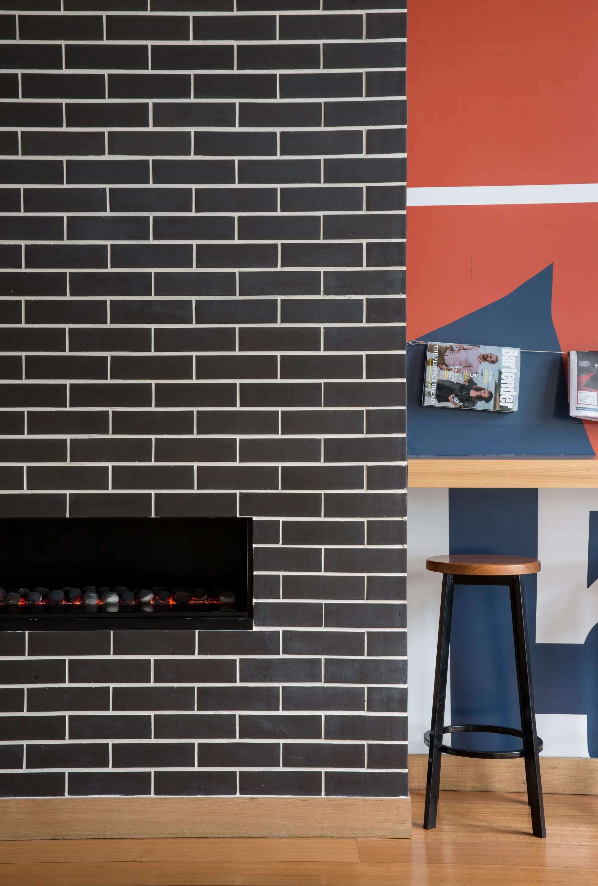 This space features a black brick fireplace with white grout and a narrow mantel displaying magazines. A wooden bar stool sits nearby against a wall decorated with bold red, white, and blue graphics.