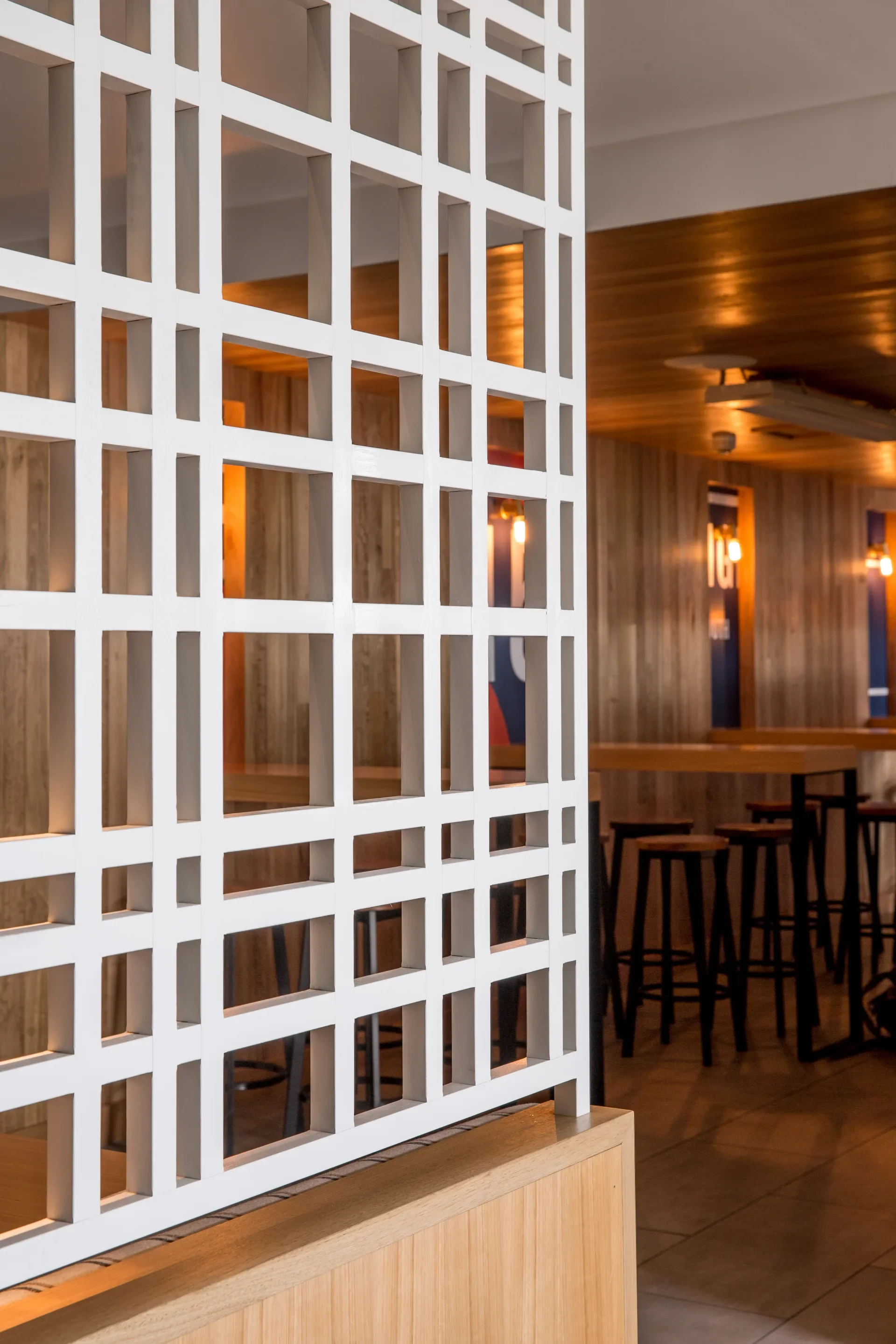 This interior view showcases a white geometric lattice screen that serves as a decorative room divider. In the background, a warm, wood-paneled space is visible, featuring high bar tables and stools illuminated by soft, glowing wall lights.