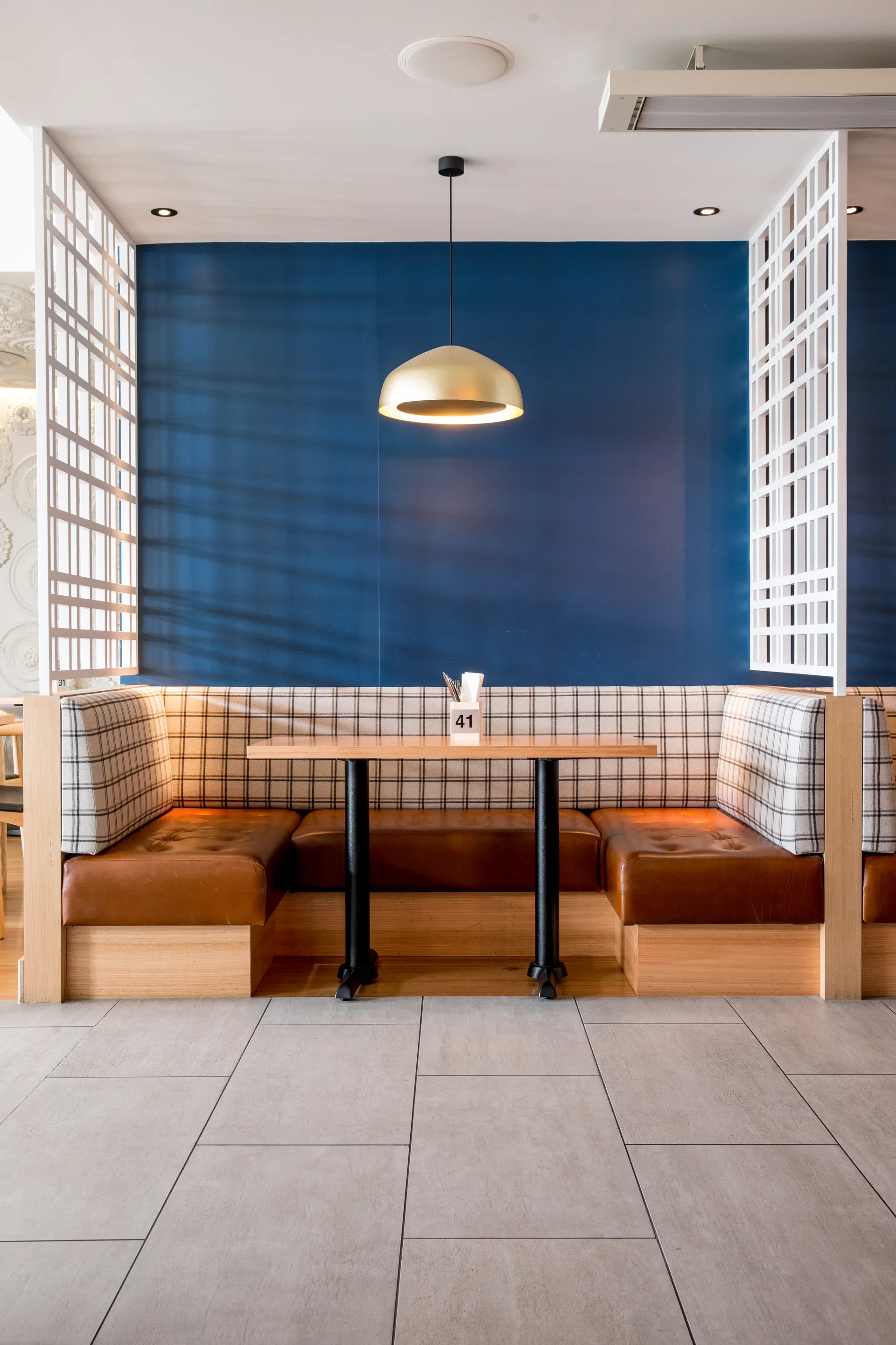 This view features a plaid-patterned booth with brown leather seating, centered against a deep blue wall. A brass pendant light hangs above the wooden table, which is flanked by white geometric lattice screens on a light tiled floor.