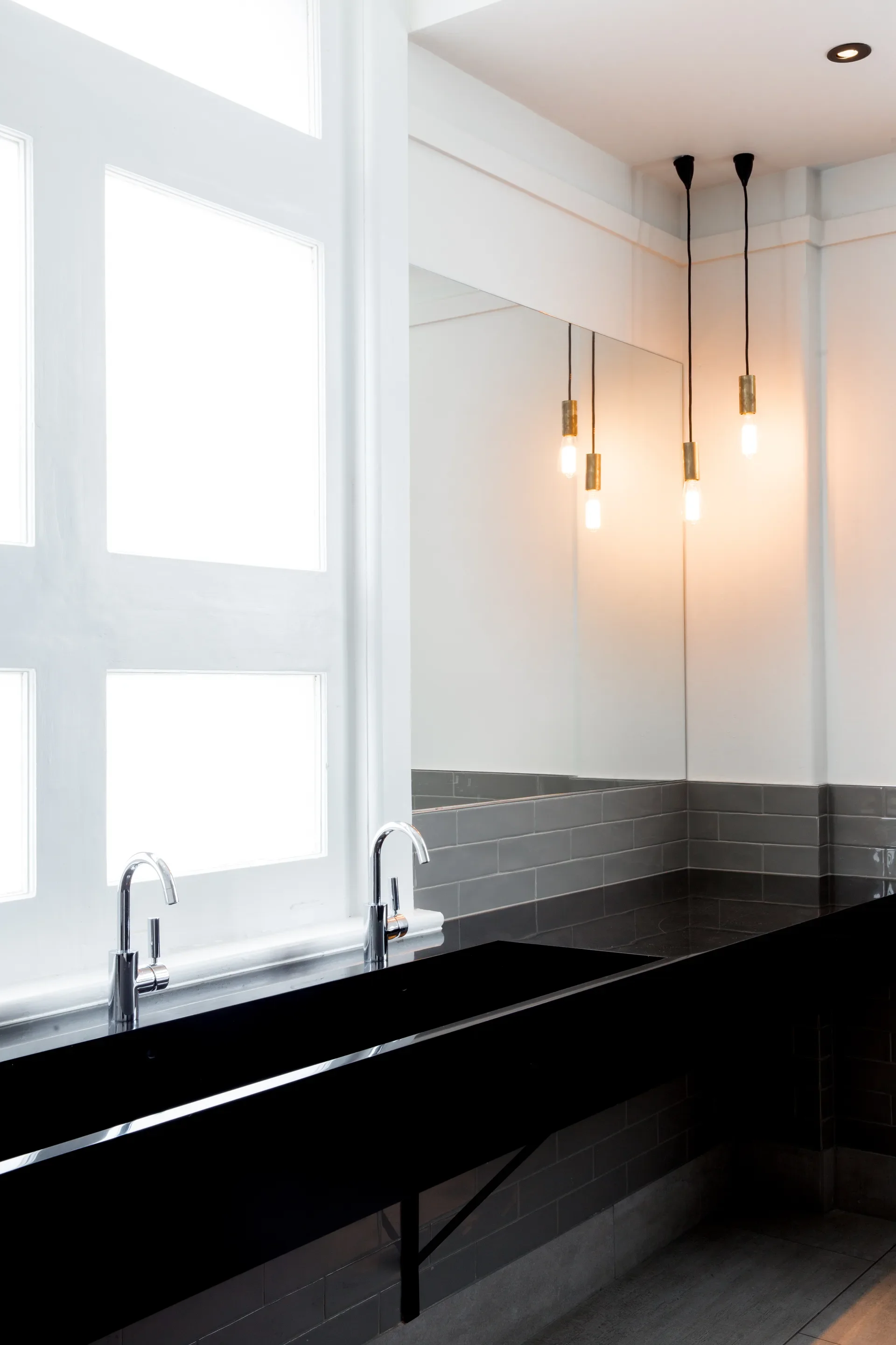 This modern bathroom features a long black trough sink with two chrome faucets set against a large white-framed window. Grey subway tiles line the wall beneath a large mirror, which reflects three minimalist pendant lights hanging from the ceiling.