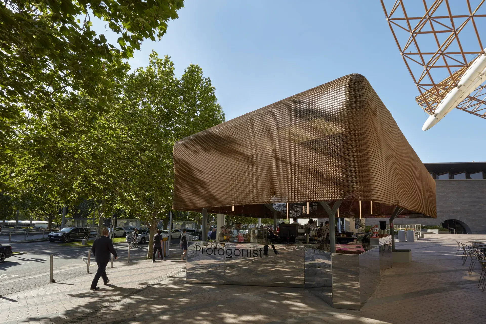 In a sunlit plaza, a rectangular bronze-mesh pavilion sits near a row of leafy green trees. People walk past the structure on a paved walkway, while a geometric metal framework and outdoor cafe seating are visible nearby.