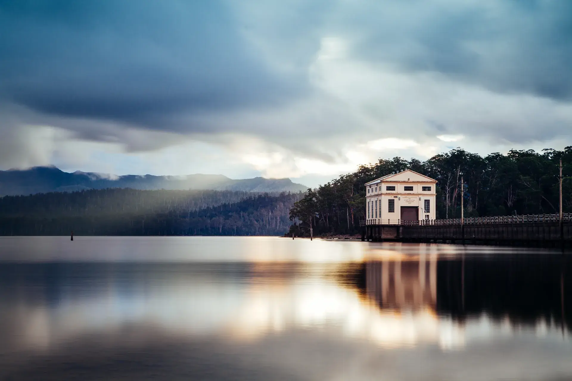 A white pump house stands at the edge of a calm lake, its reflection shimmering on the water's surface. Behind it, a dense forest covers the rolling hills under a dramatic, cloud-filled sky.