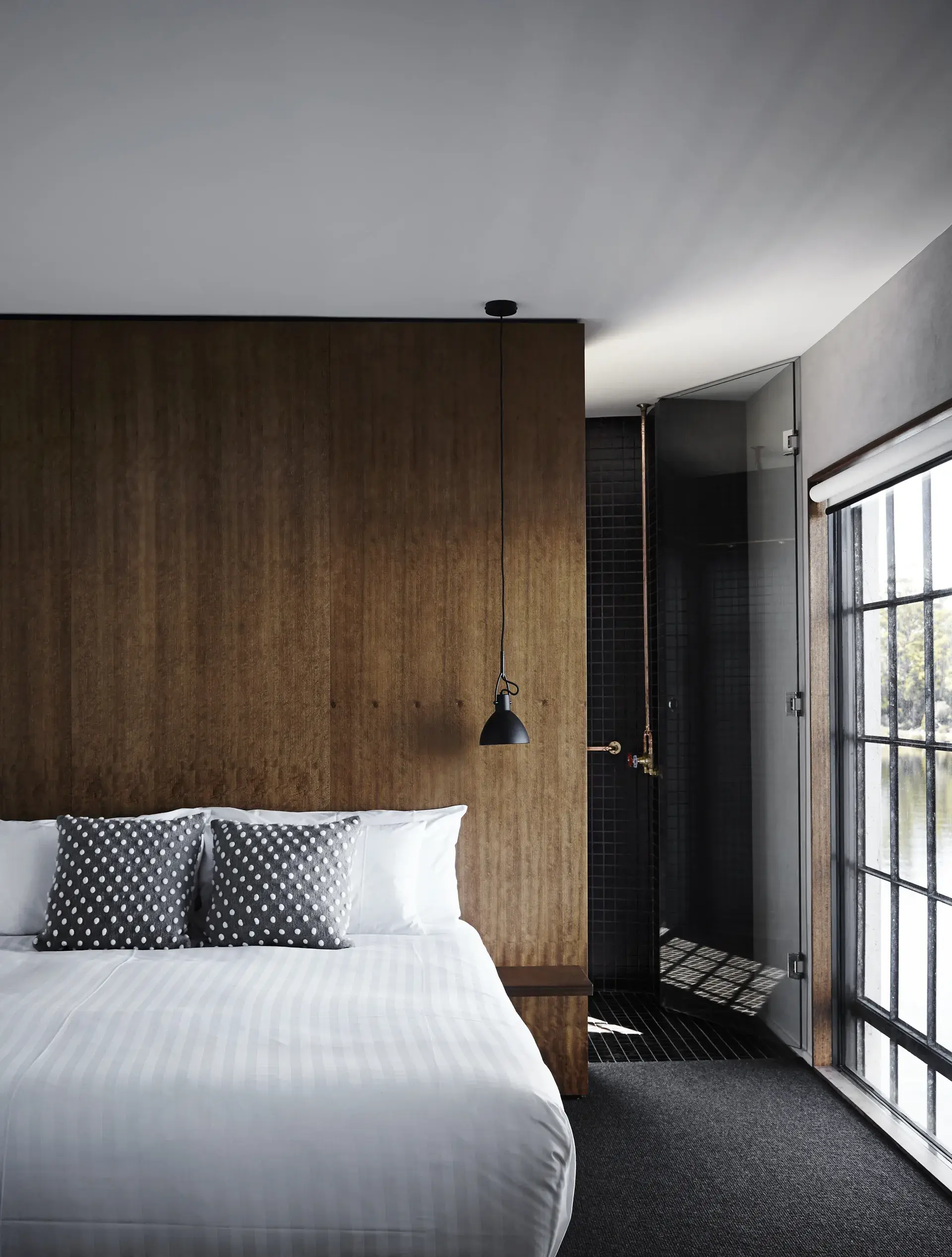This modern bedroom features a wood-panelled wall and a bed with white striped linens and grey polka-dot pillows. An industrial-style window overlooks the water, and a black-tiled shower is visible through a glass door.