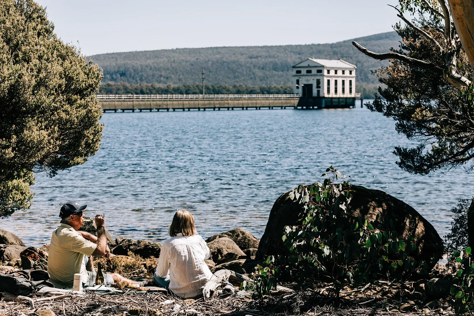 Two people sit on a rocky shoreline, enjoying a picnic with a view of a white pumphouse and its long pier across the lake. The scene is framed by native green shrubs and a large boulder, with forested hills visible under a clear, bright sky.