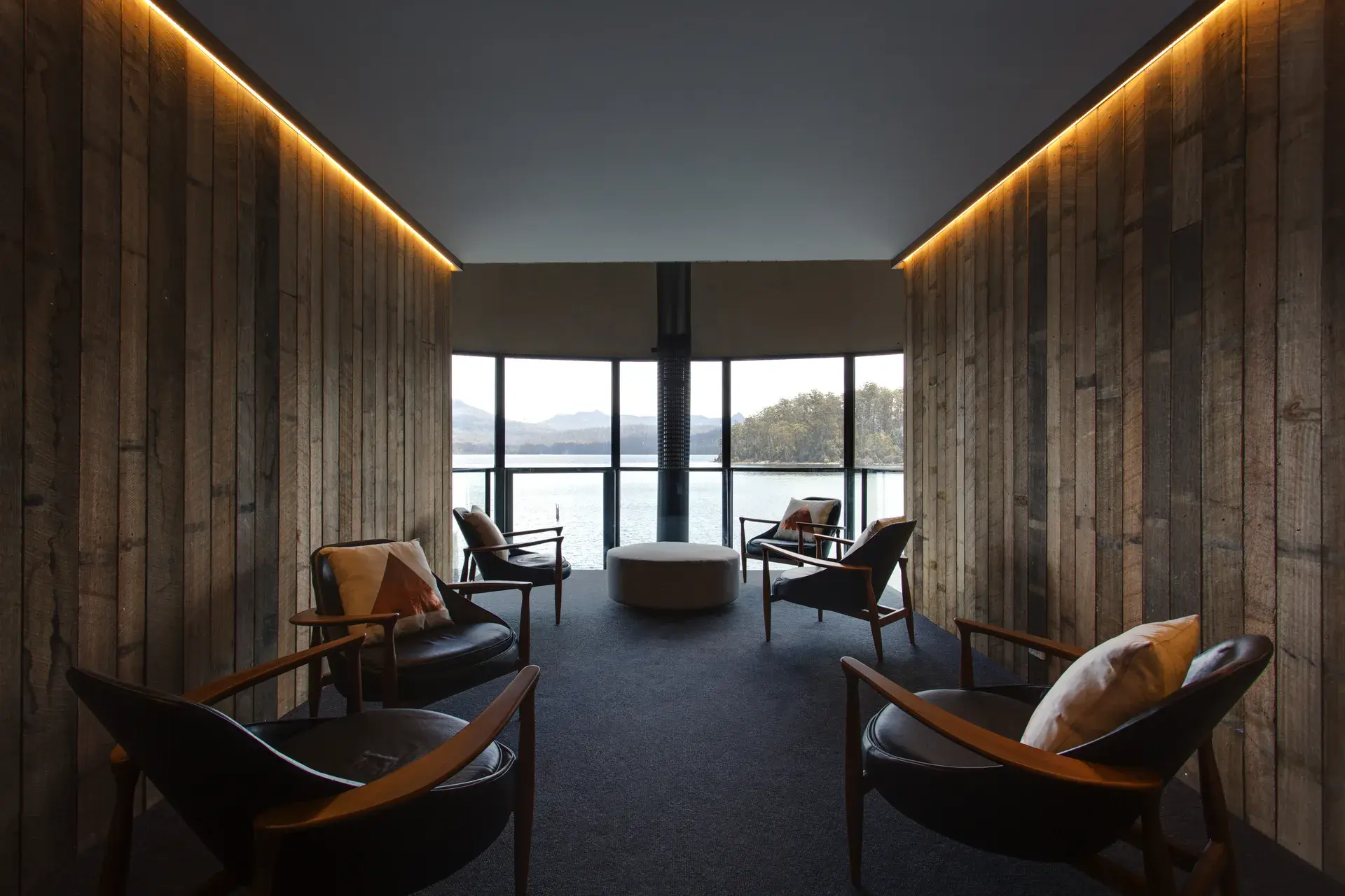 This lounge features vertical wooden slat walls with warm, integrated strip lighting along the ceiling. Several dark leather armchairs are arranged around a central ottoman, all facing a large window that offers a panoramic view of a lake and forested islands.