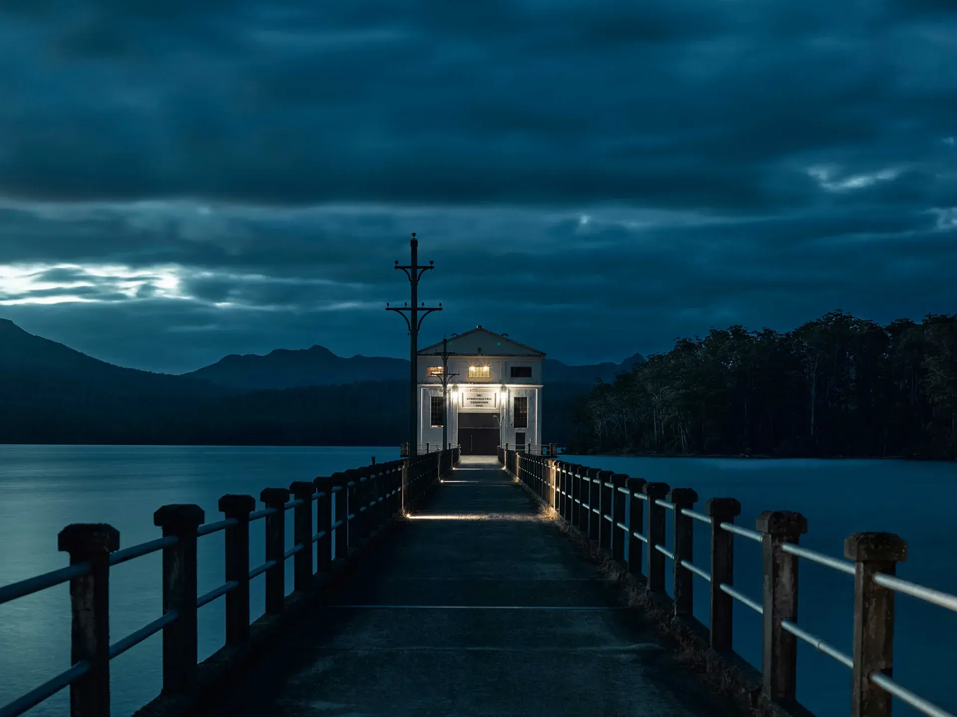 This night view shows a long concrete pier leading to a white pumphouse at the edge of a lake. Warm lights illuminate the walkway and the building's facade against a backdrop of dark mountains and a moody, blue-toned sky.