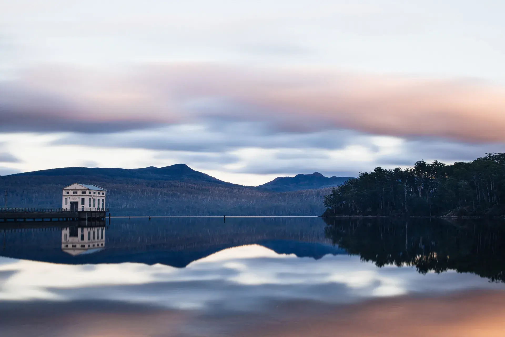 A white pump house stands at the edge of a calm lake, its reflection shimmering on the water's surface. Behind it, a dense forest covers the rolling hills under a dramatic, cloud-filled sky.