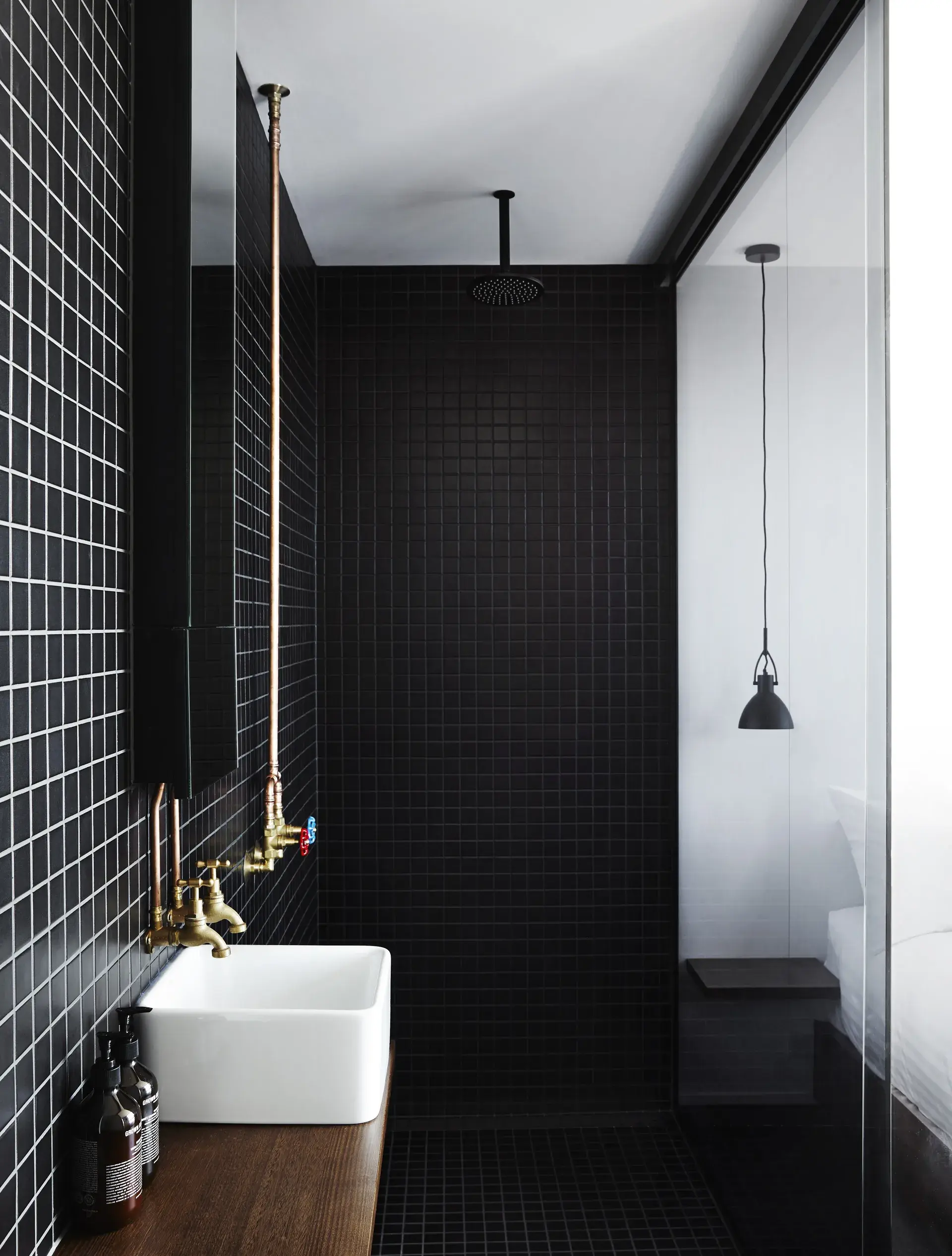This modern bathroom features matte black square tiles on the walls and floor, contrasted by a white vessel sink on a wooden counter. Exposed copper pipes and brass fixtures add an industrial feel, while a glass partition separates the shower from the bedroom.