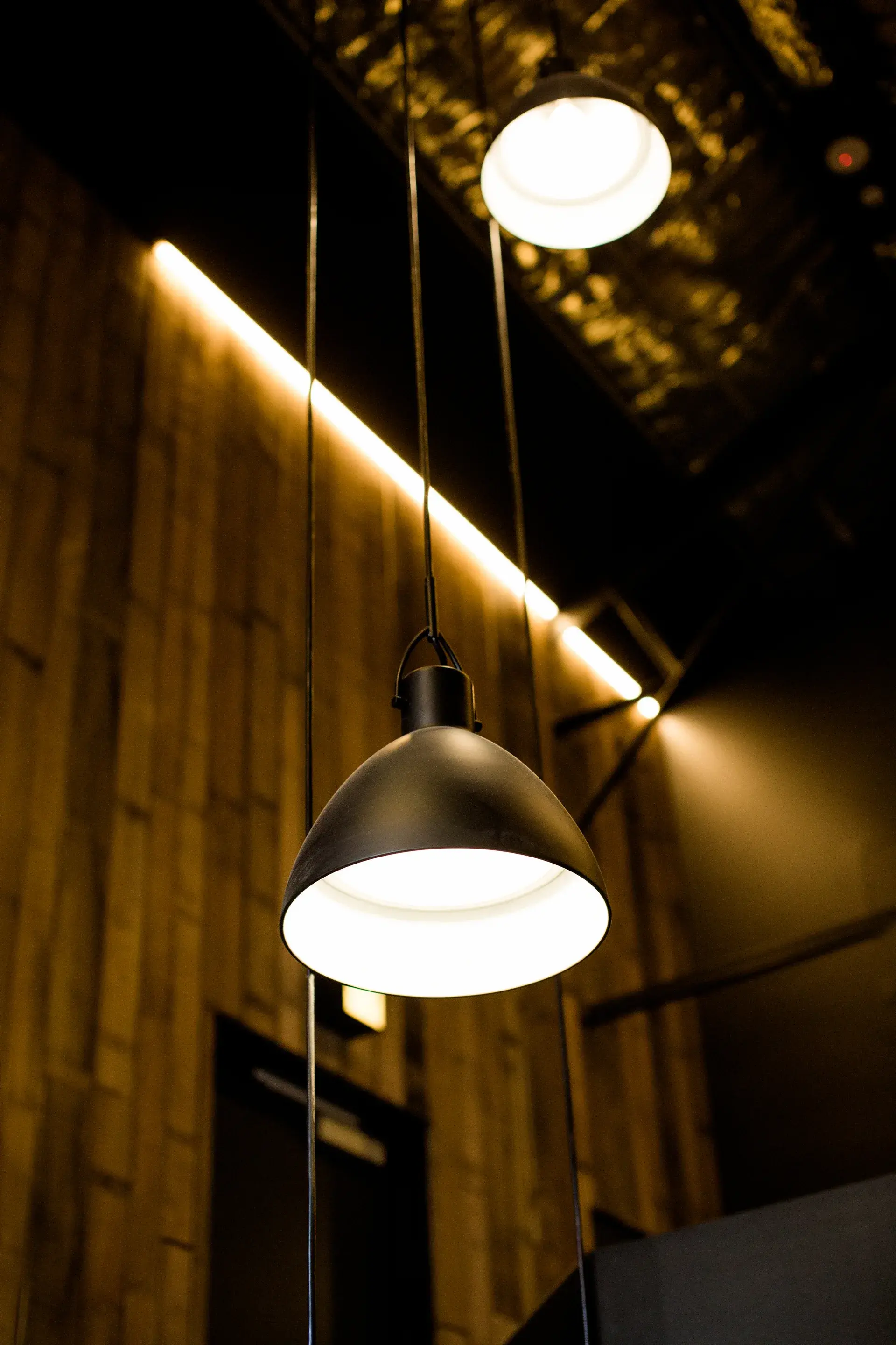 This interior features black dome pendant lights hanging from the ceiling, casting a warm glow. In the background, vertical wood-panelled walls are accented by a bright, horizontal LED light strip.
