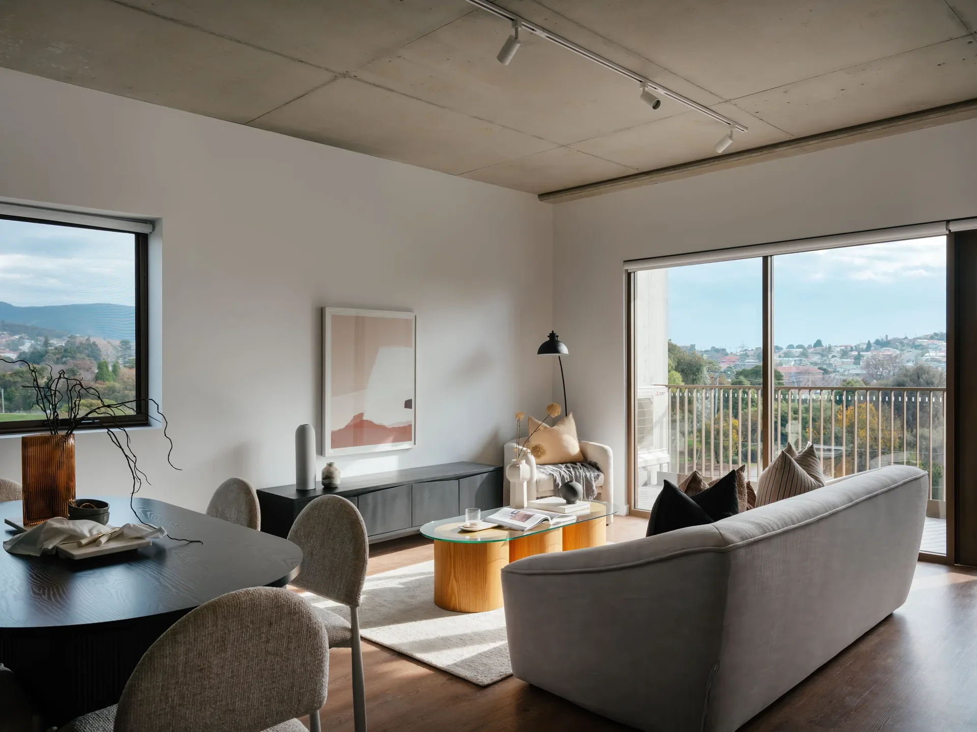 The living and dining area features an exposed concrete ceiling with white track lighting. A light grey sofa and wooden coffee table sit near a large glass door that opens onto a balcony overlooking the city.