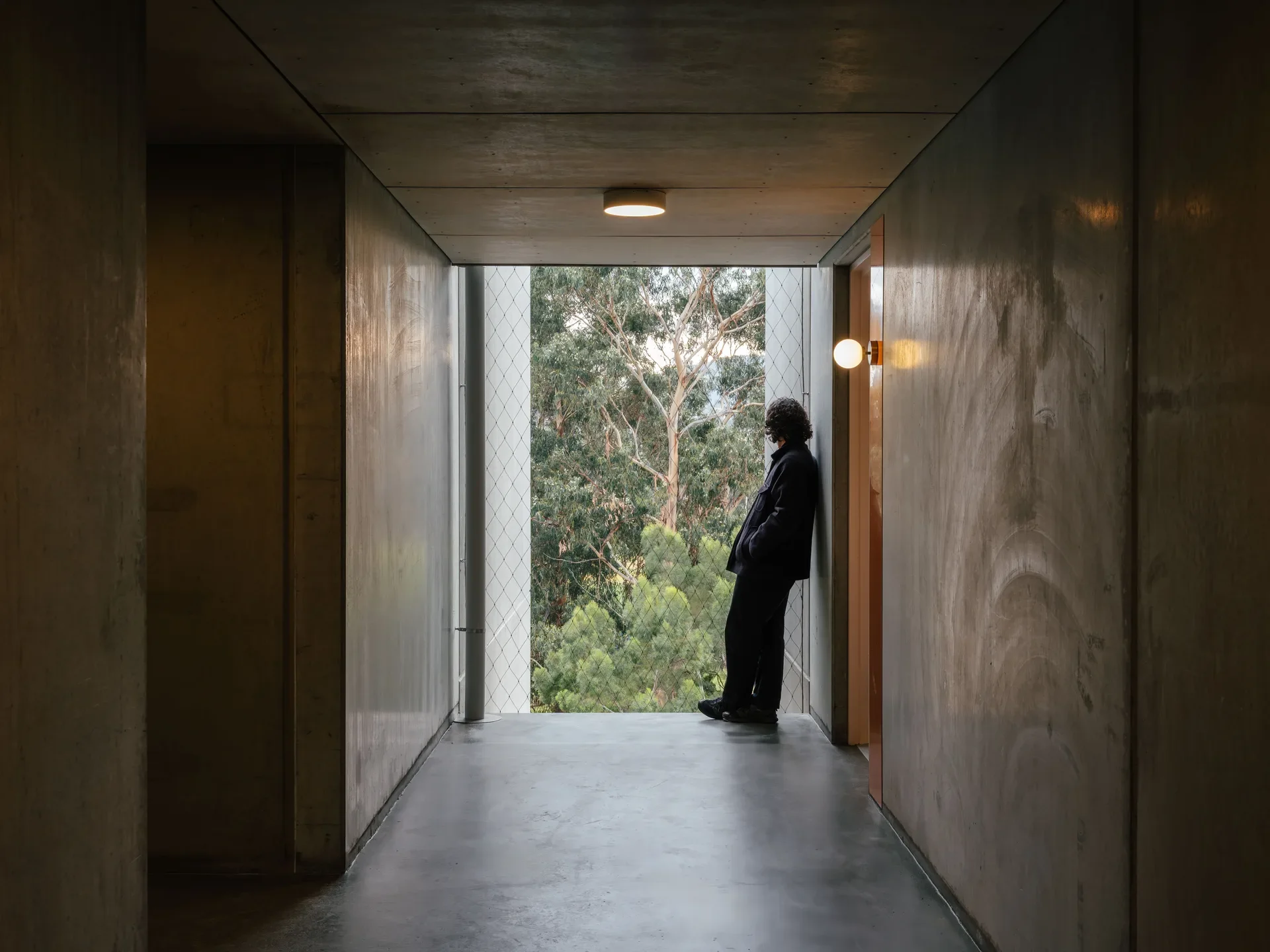This industrial hallway features exposed concrete walls and flooring which leads to an opening with a wire safety net. A person stands looking out at a dense green forest.