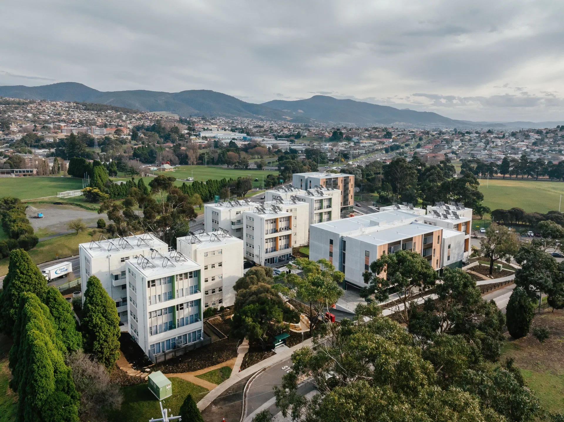 This aerial view shows a residential complex consisting of several buildings buildings, two of which are additions to the existing site. The structures are topped with solar panels and are surrounded by green lawns, trees, and a distant mountain range under a cloudy sky.
