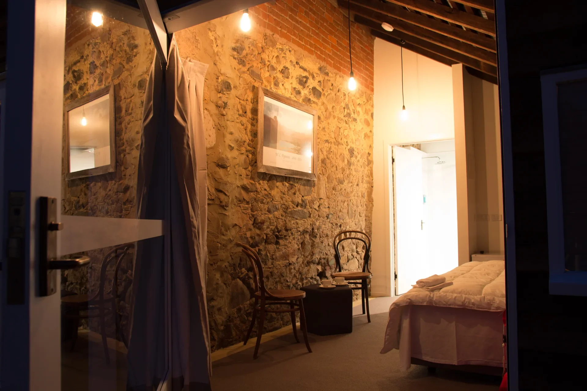 This rustic bedroom features an exposed stone wall and a slanted ceiling with wooden beams. Warm pendant lights illuminate a bed with white linens and a small seating area with two wooden chairs.