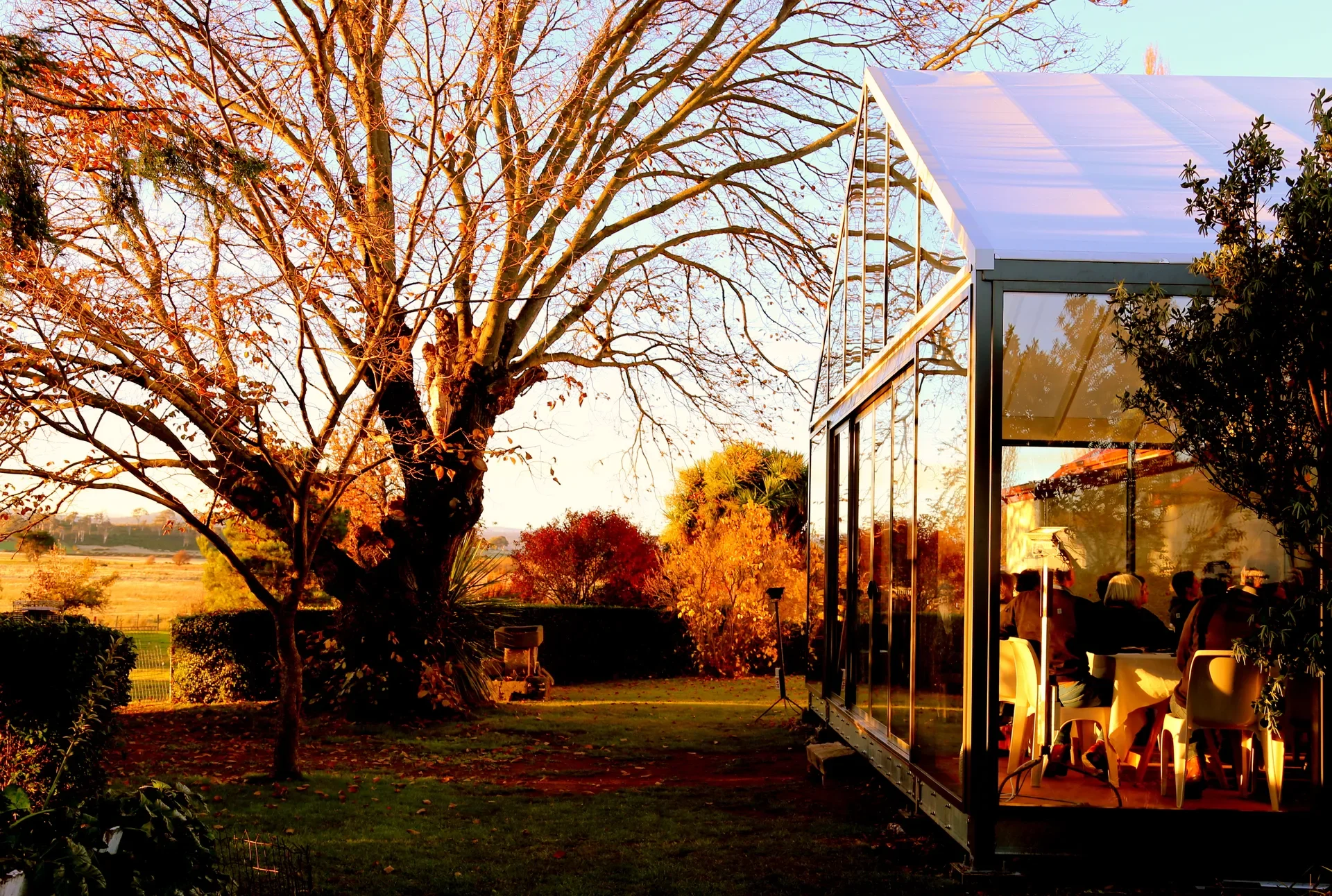 A glass-walled structure with a gabled roof stands in a garden, with people visible dining inside. Outside, large trees with autumnal orange leaves frame a view of a distant field under a warm, golden light.
