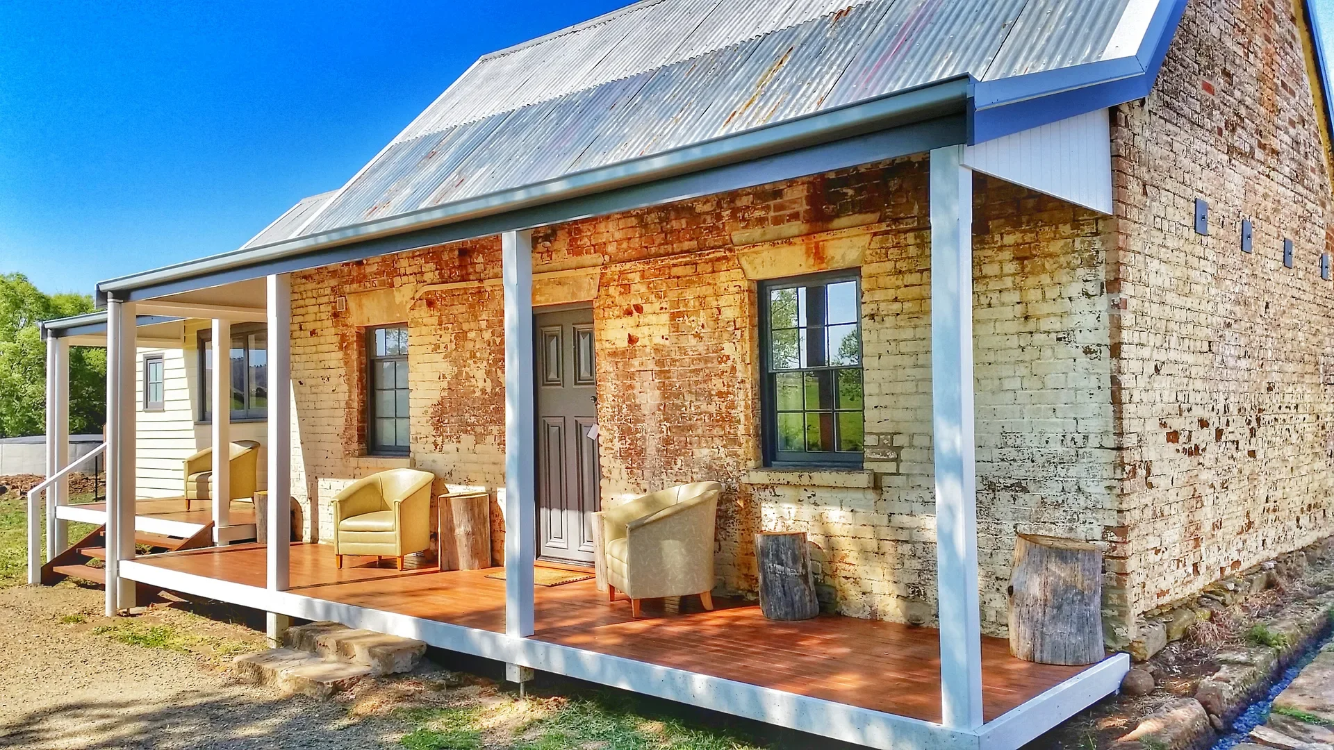 This weathered brick cottage features a long wooden porch with white support beams and several tan armchairs. The building is topped with a corrugated metal roof, and a small stone drainage channel runs alongside its base.