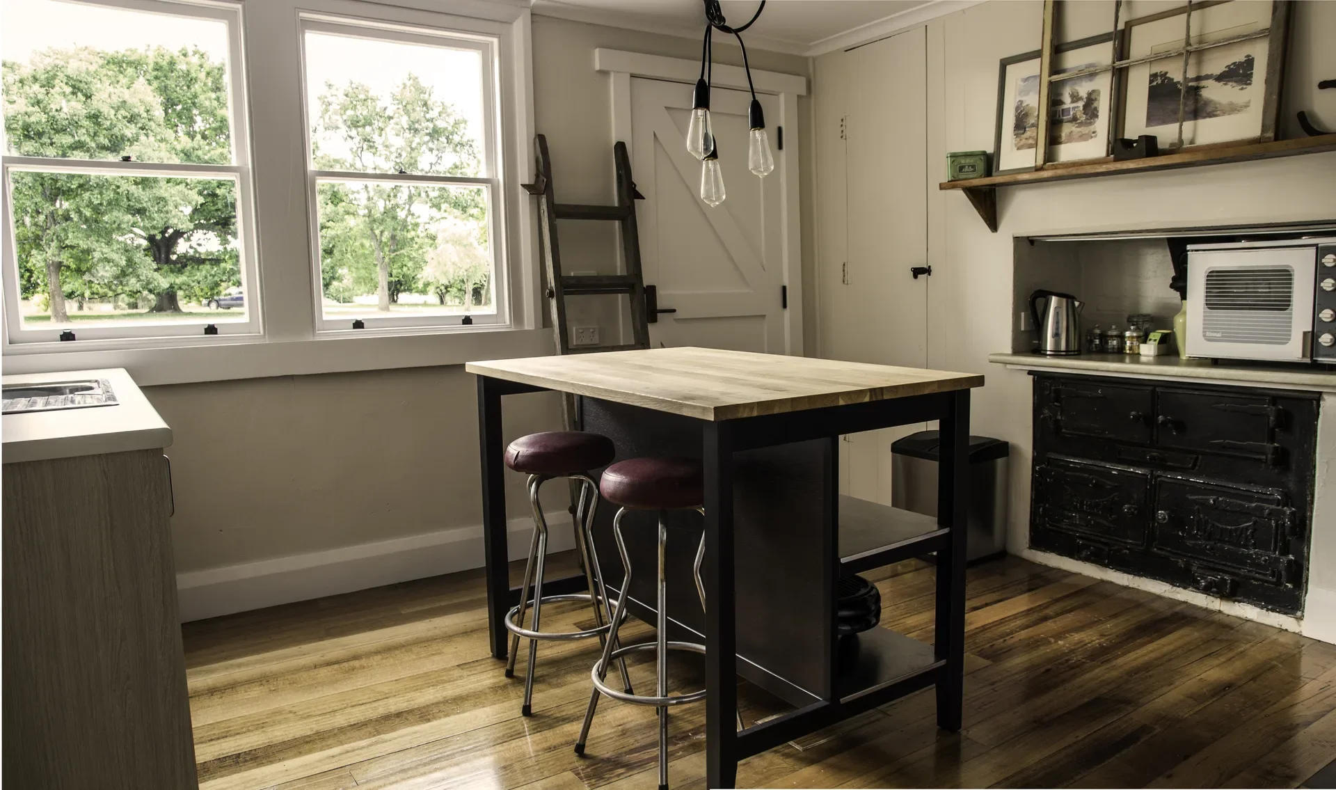 This rustic kitchen features a wooden island table with two metal barstools and an old-fashioned black wood-burning stove. Large windows look out onto a green yard, while a wooden ladder and Edison-style pendant lights add to the vintage aesthetic.