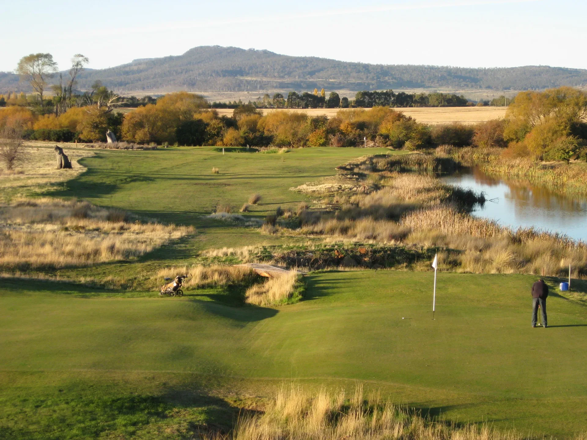 A golfer stands on a green next to a white flag, overlooking a expansive golf course. The landscape features a winding waterway lined with tall grass, leading toward rolling hills and distant mountains under a clear sky.
