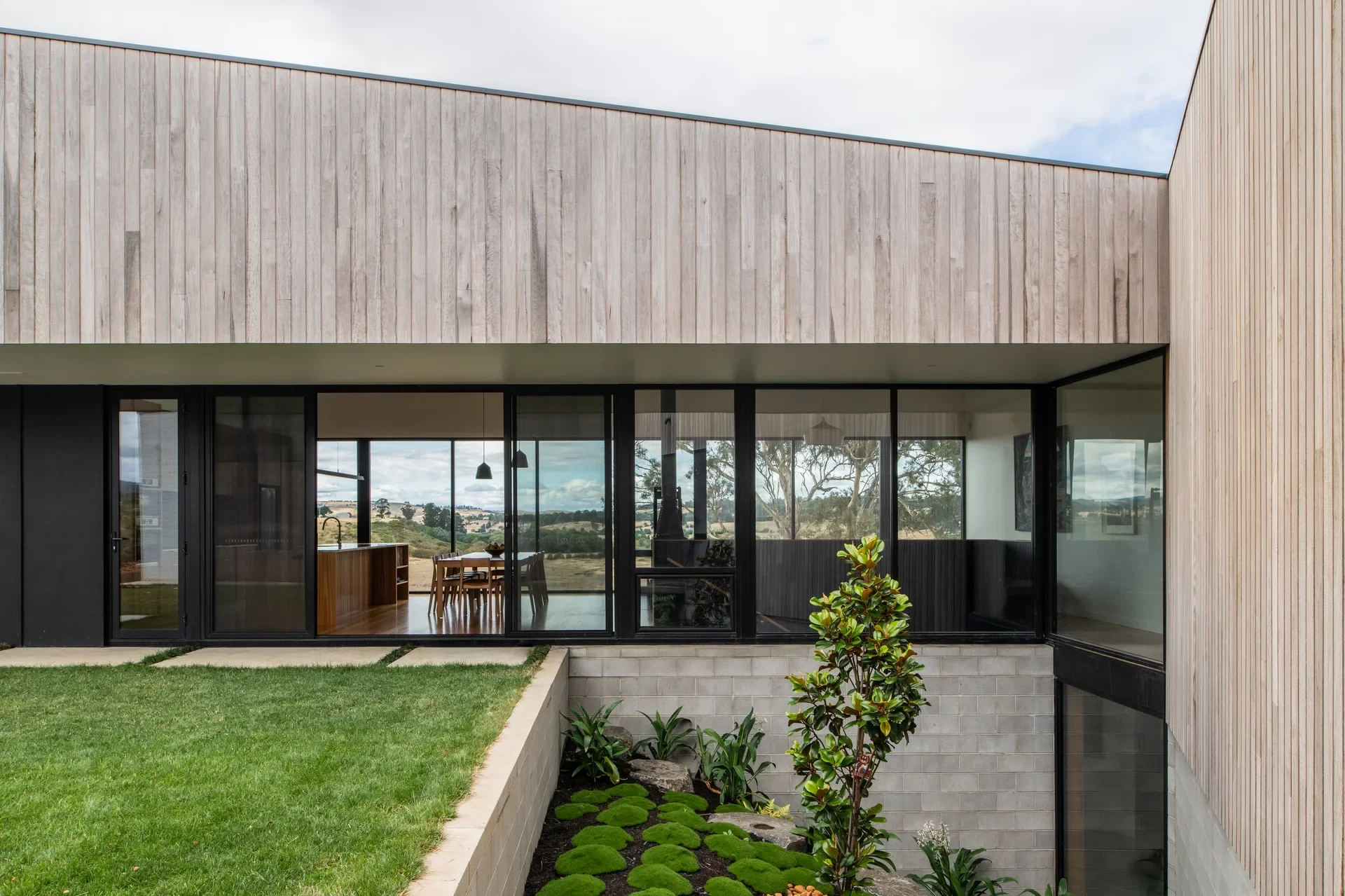 A timber-clad home with large black-framed windows overlooking a rural landscape. Below the lawn, a sunken garden with mossy mounds and structural plants adds a tiered, architectural element.