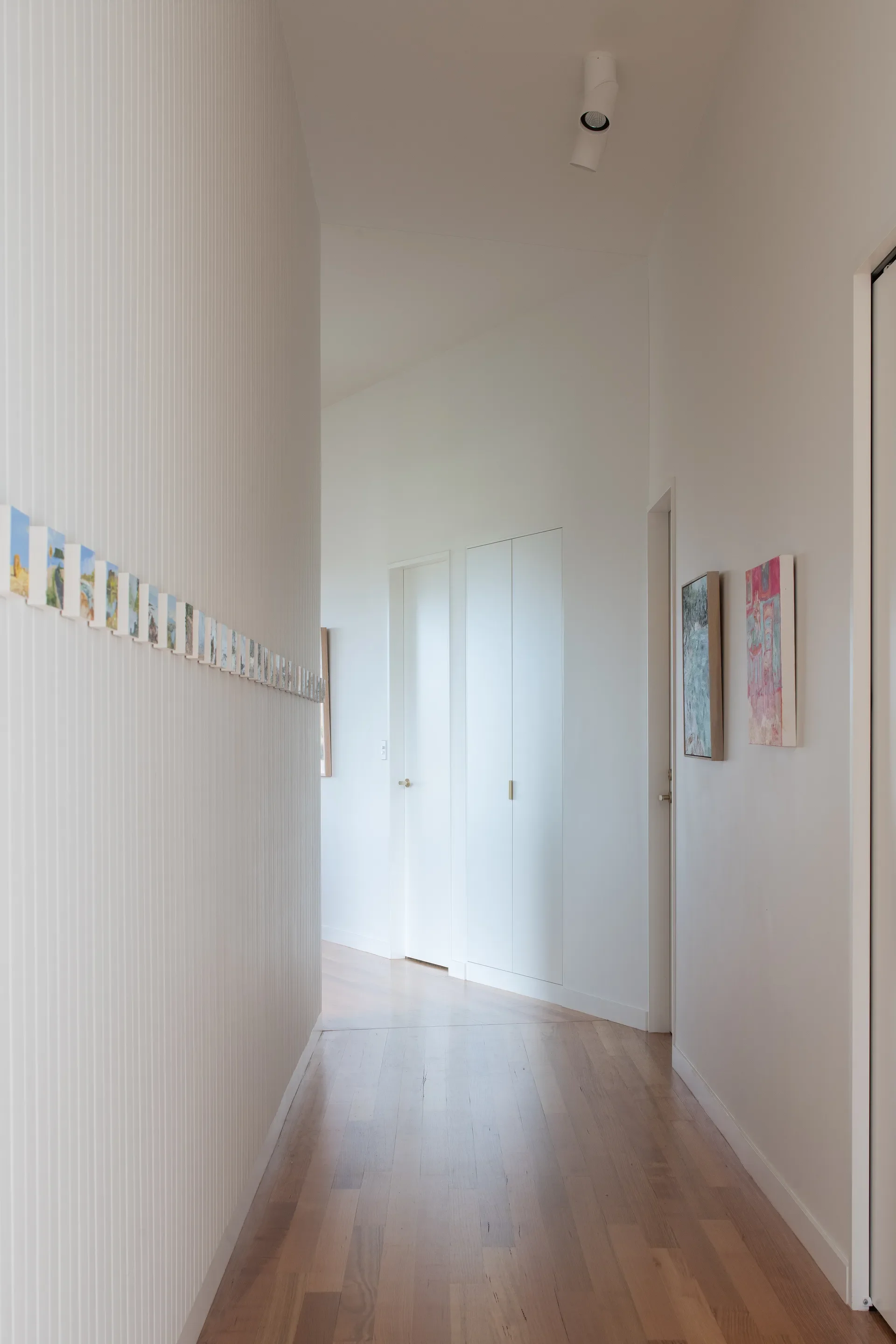 Minimalist white hallway featuring vertical-slat wall paneling and light timber flooring. A long, linear series of small artworks lines the left wall, while vibrant framed paintings add a splash of colour to the right.