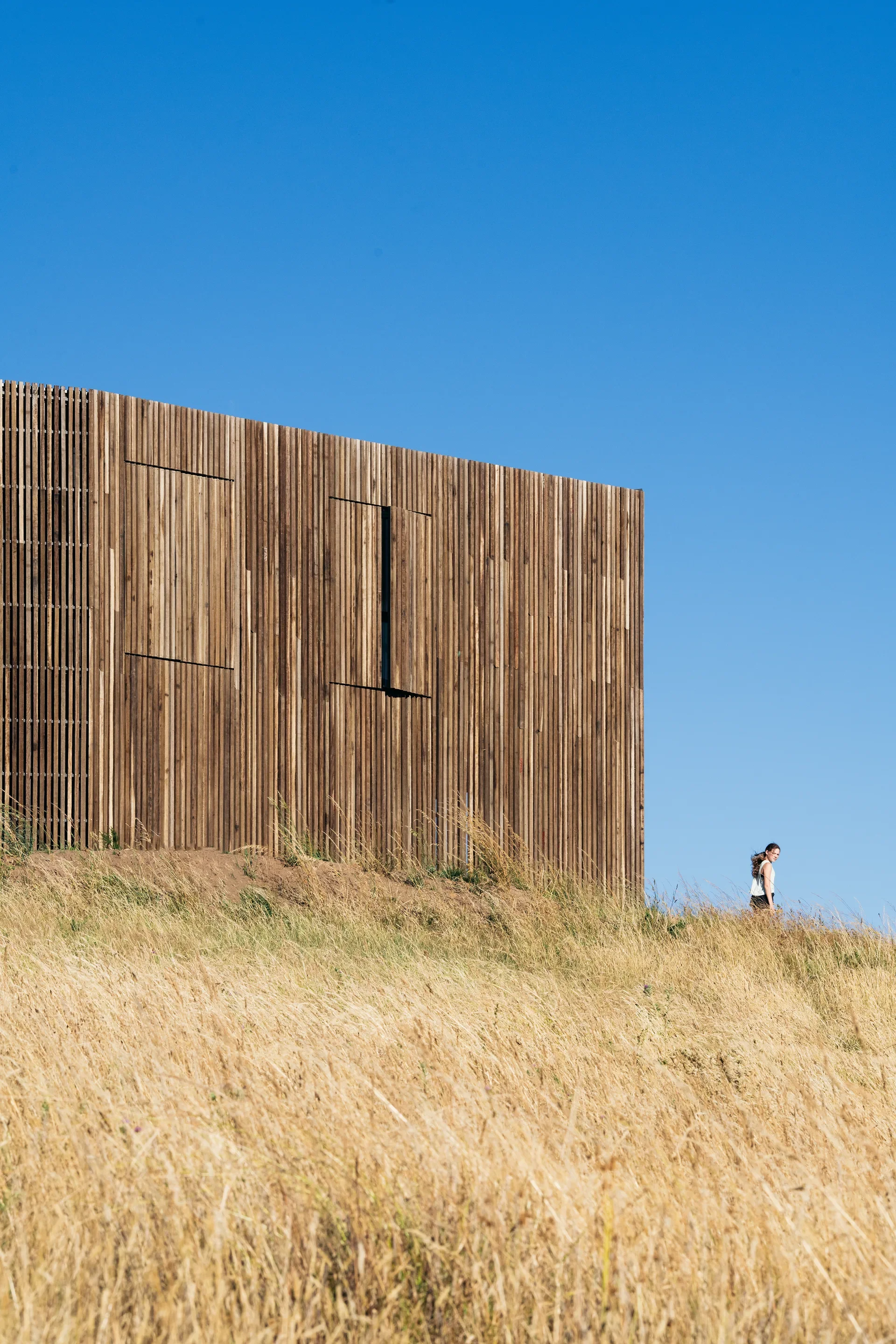 Modern timber-slat building with a tall, rectangular profile standing on a golden, grassy ridge. A figure walks along the hillside under a clear, vibrant blue sky, highlighting the contrast between the organic wood texture and the minimalist horizon.