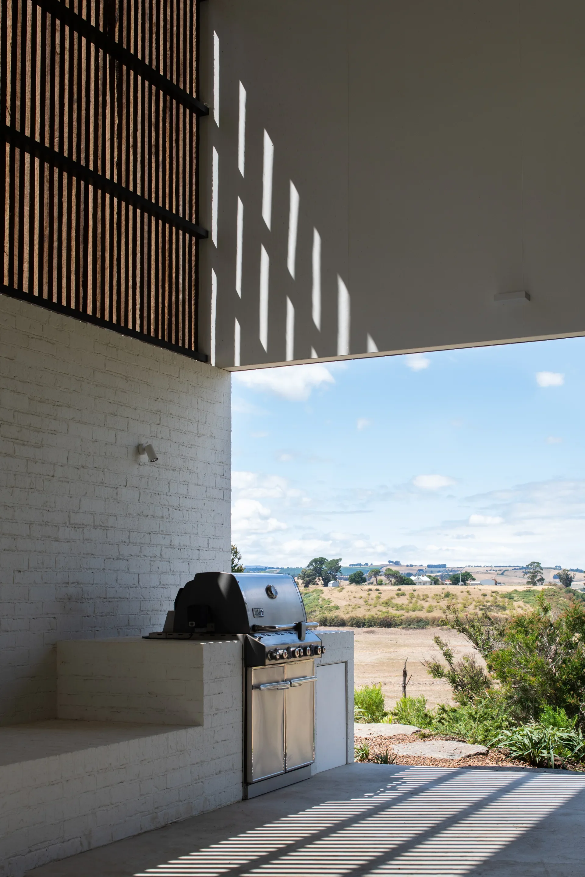 Modern outdoor kitchen with a built-in barbecue set against white brick walls. Sunlight filters through a slatted timber screen, casting geometric shadows over the patio.