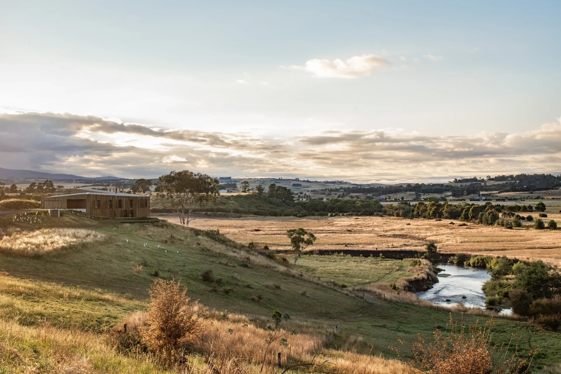 Panoramic view of a timber-clad house nestled on a golden ridge overlooking a winding river. The vast, sunlit landscape stretches toward rolling hills under a soft, cloudy horizon.