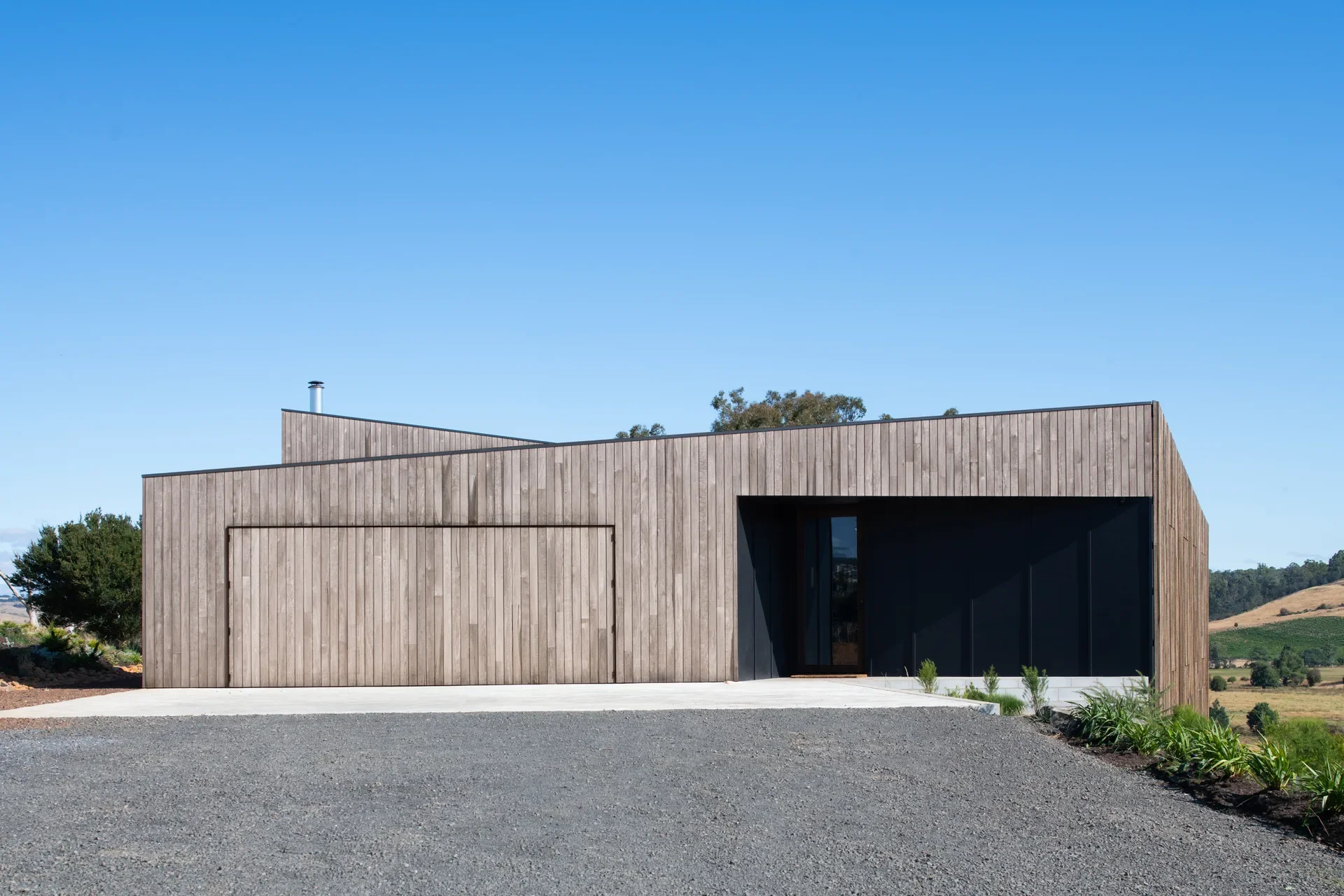 A weathered timber home with sharp, geometric rooflines and a hidden garage door. The minimalist structure is set against a gravel driveway and a clear blue sky, overlooking a rural valley.