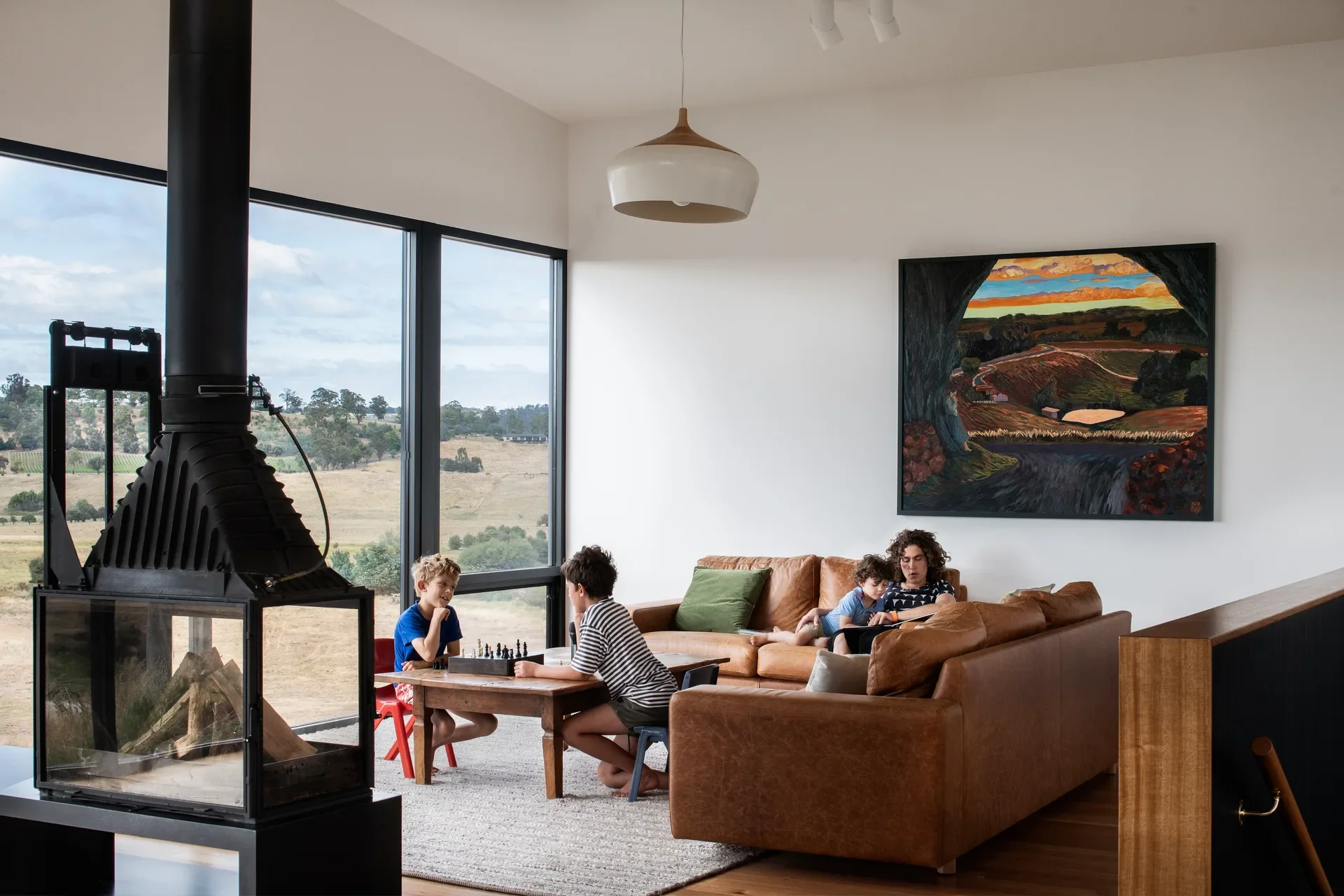 Sun-drenched modern living room featuring a family relaxing by a wood-burning fireplace. Large windows and a landscape painting mirror the rolling golden valley outside.
