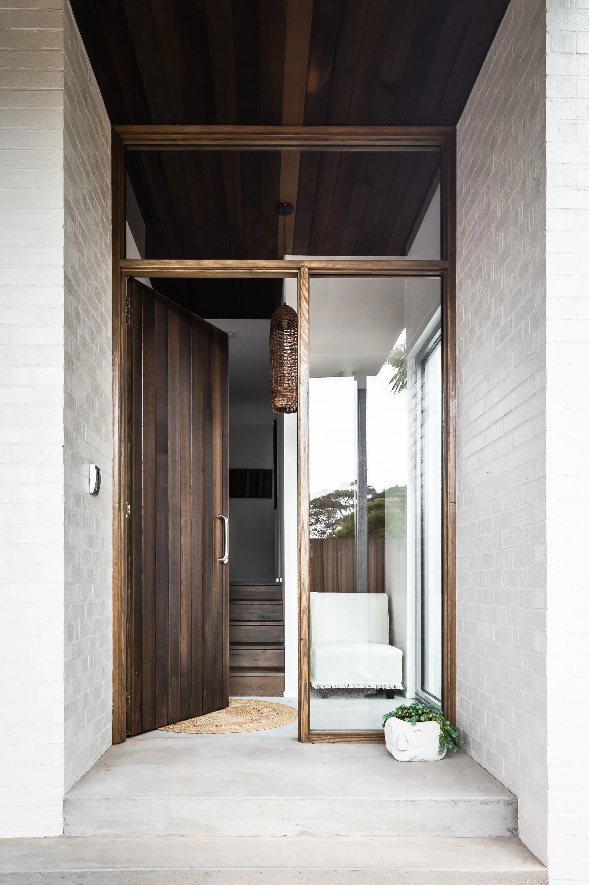 This modern entryway features a tall wooden door and a matching dark wood ceiling. Adjacent to the door, a large glass window reveals a white chair and an interior staircase, with a woven pendant light hanging above.